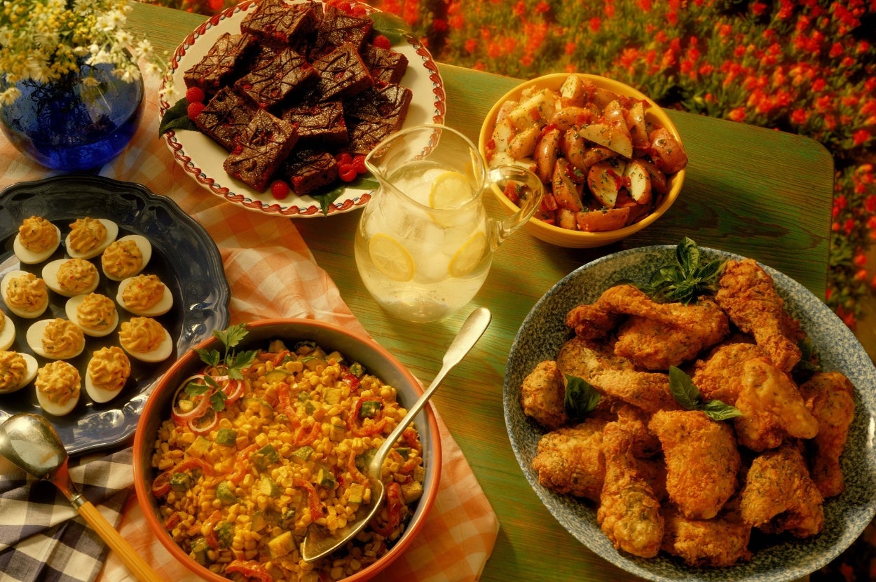 A table with fried chicken, roasted potatoes, deviled eggs, corn salad, brownies on plates, and a pitcher of lemonade