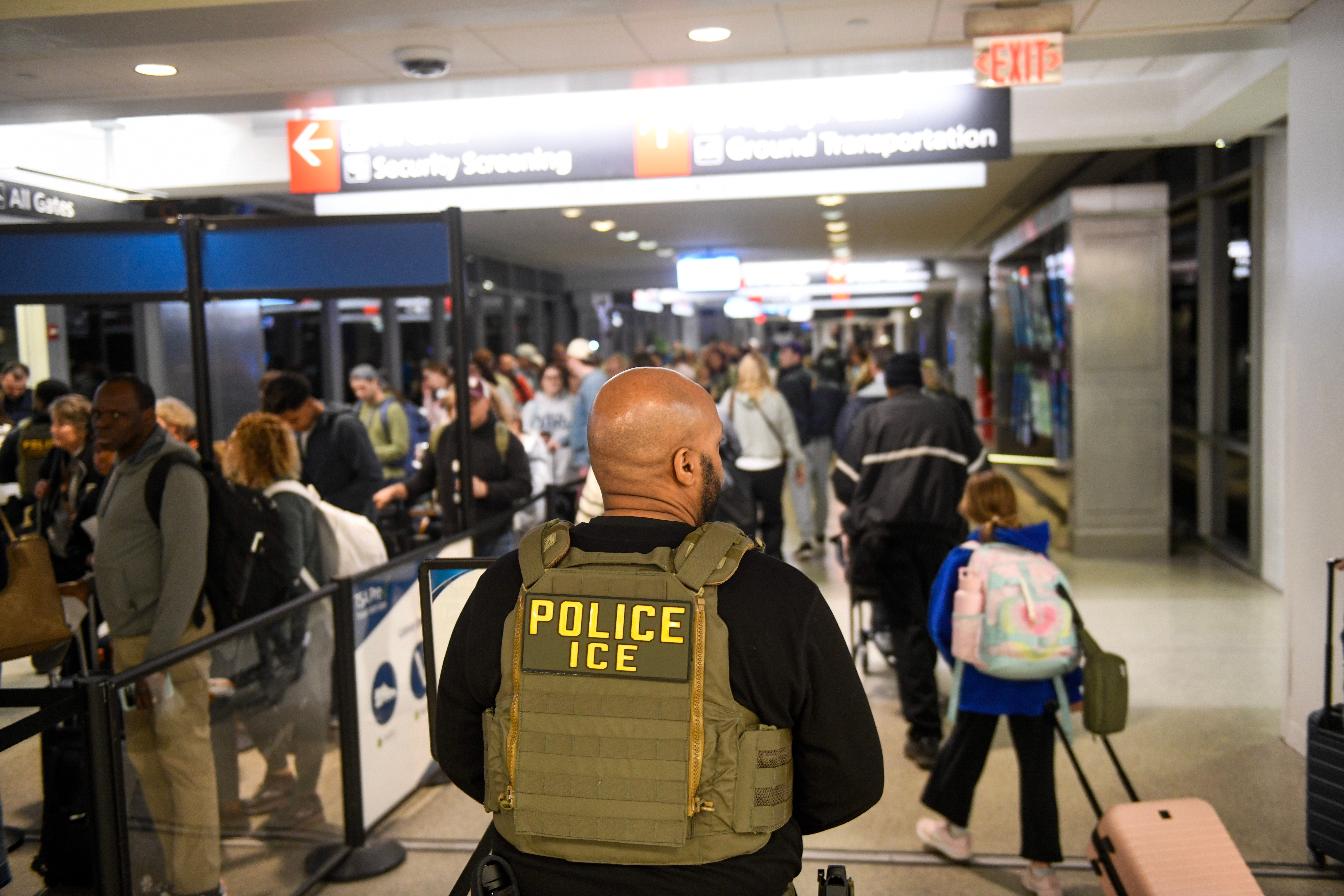 Sikkerhetsoffiser iført "Politi ICE" vest overvåker travle flyplassterminal; folk står i kø ved sikkerhetskontrollen