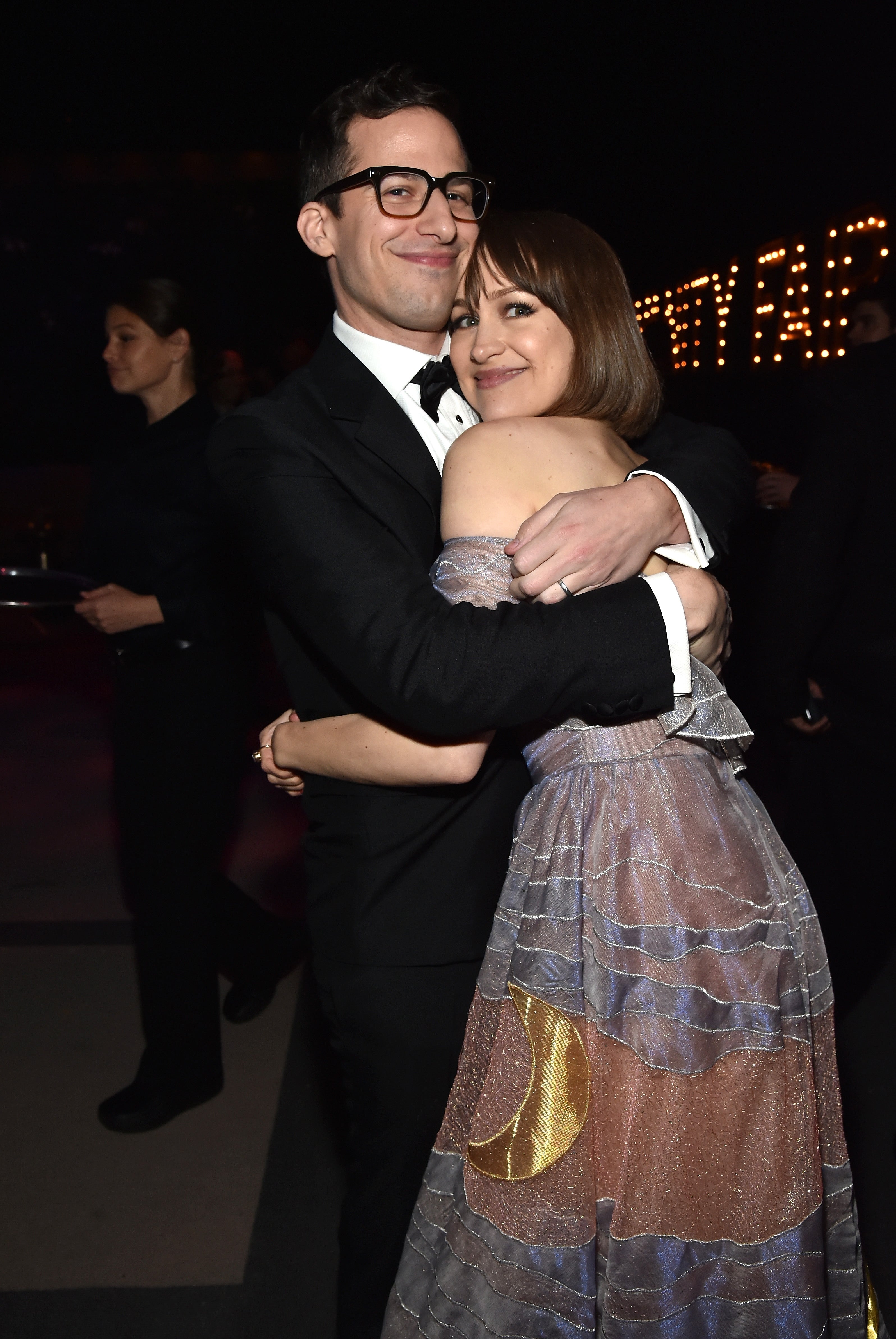 Andy Samberg and Joanna Newsom wear formal attire and embrace at an event. Newsom wears an off-shoulder dress, and Samberg wears a tuxedo