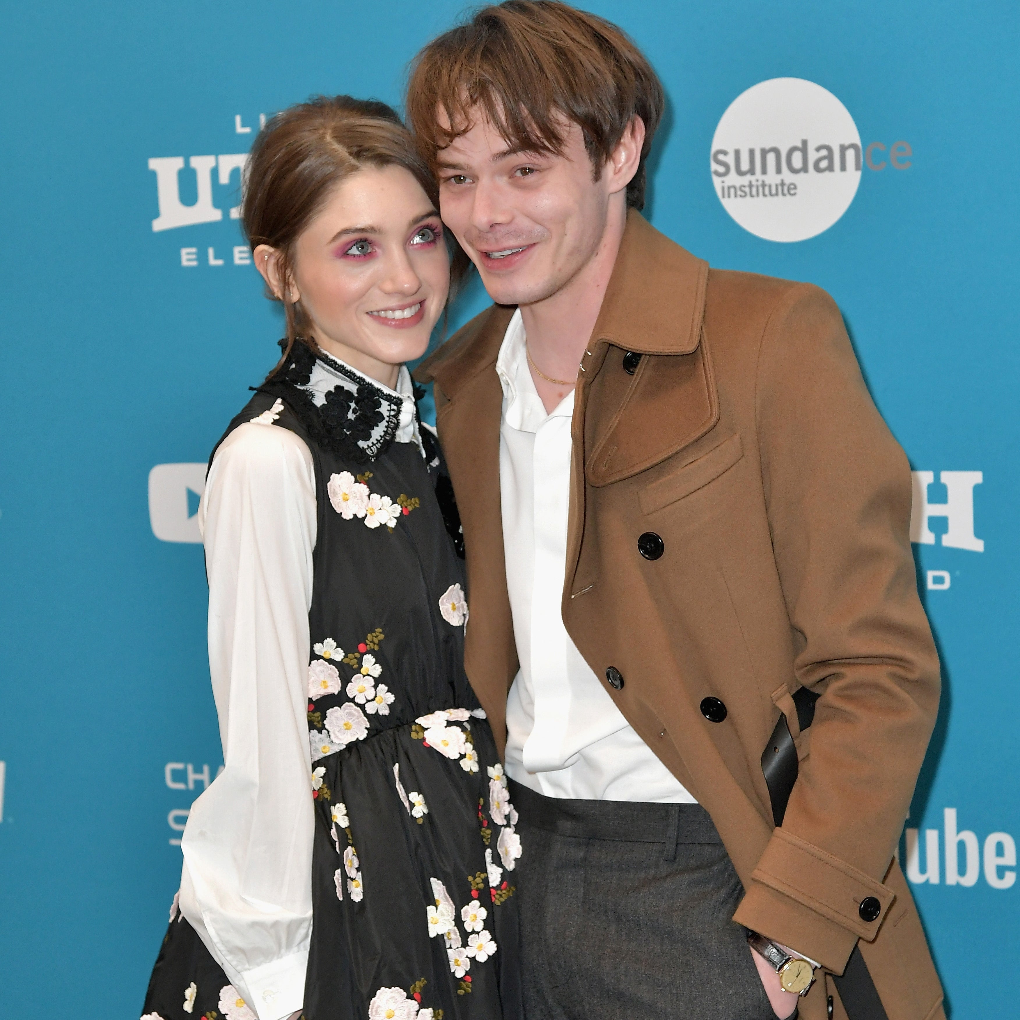 Natalia Dyer and Charlie Heaton pose at a film event. Dyer wears a floral dress with a white top, and Heaton wears a tan coat over a white shirt and gray pants