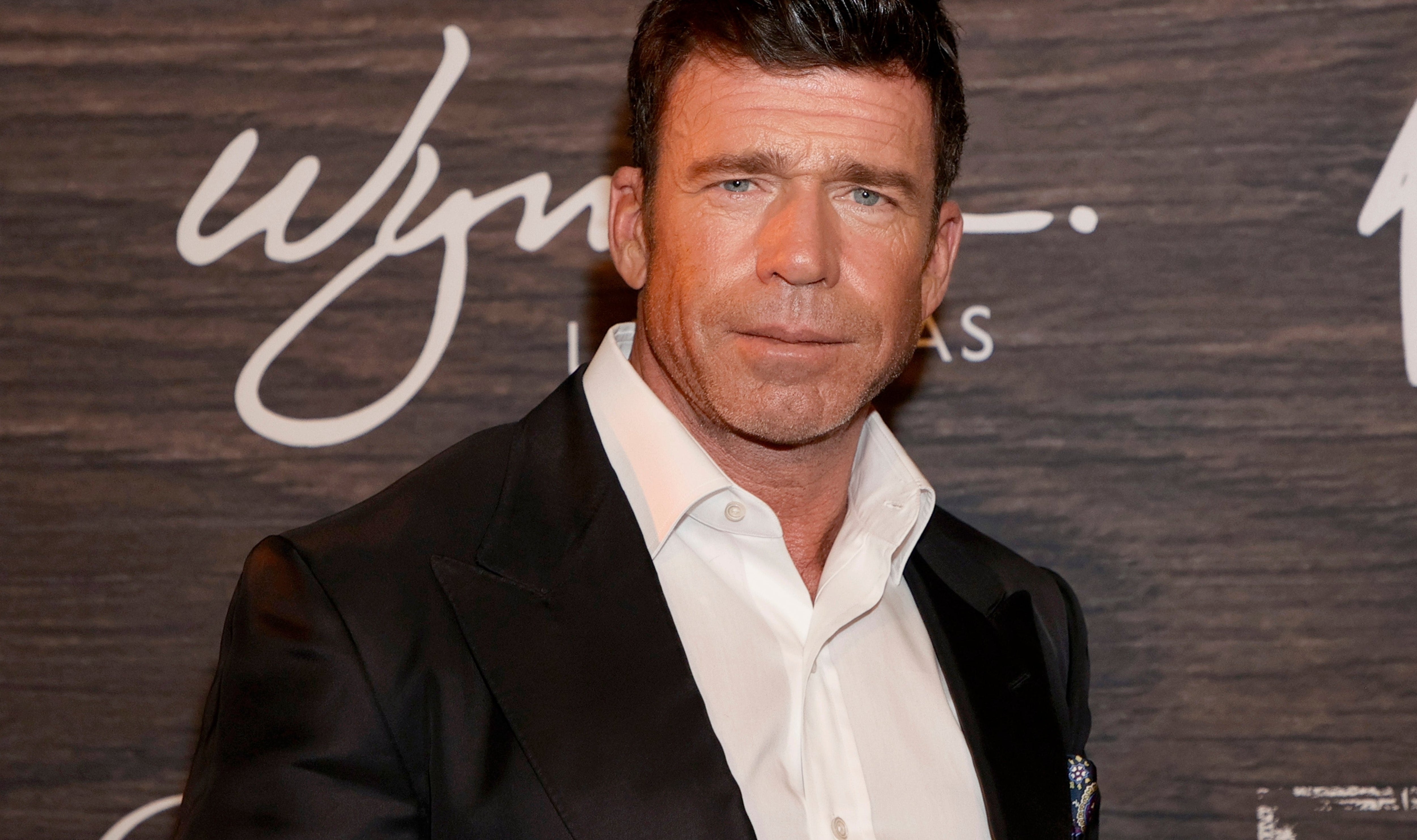 Person in formal attire at an event, wearing a white shirt and black blazer, standing against a backdrop with logo and text