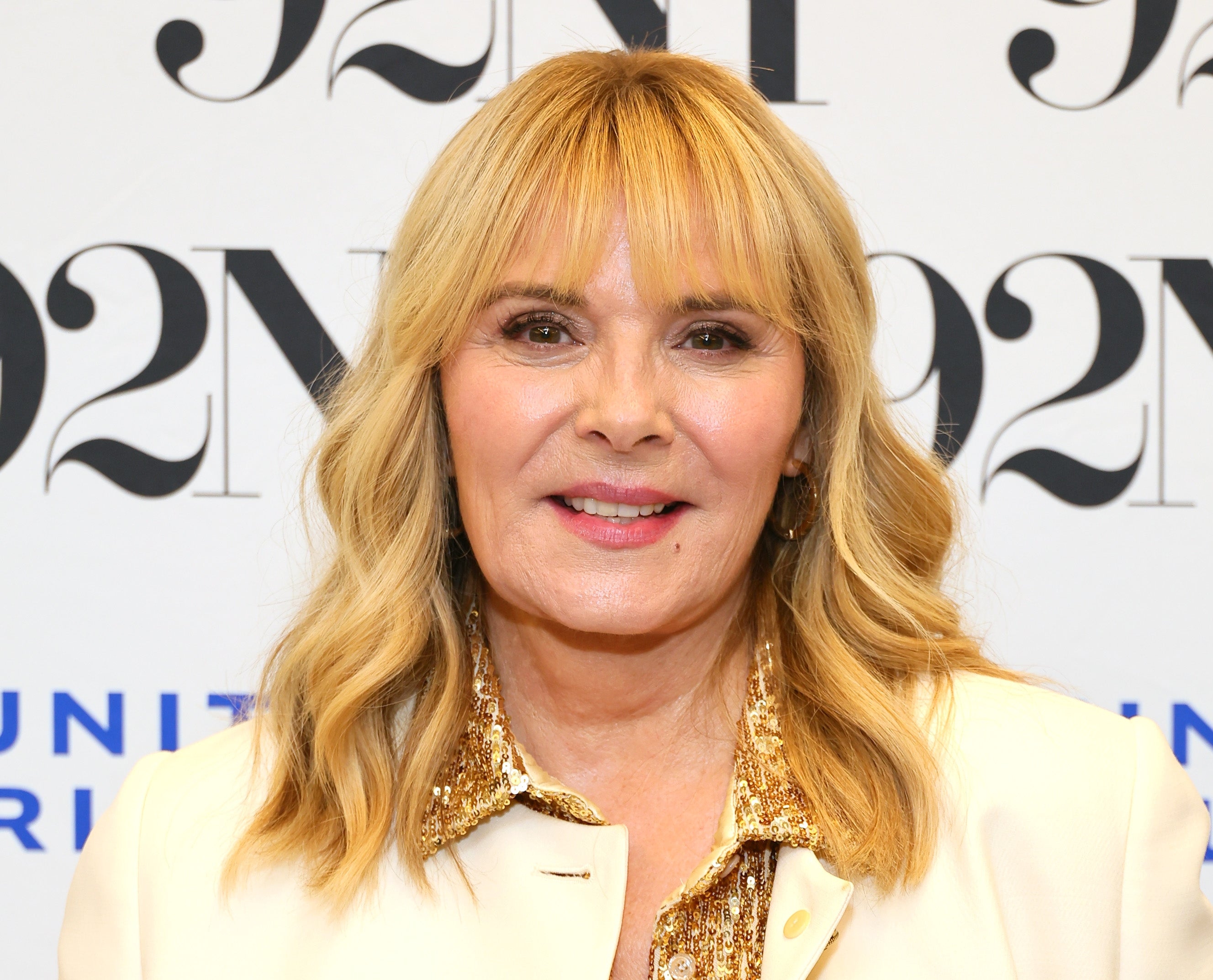 Person with wavy hair wearing a cream-colored jacket and glittery gold top, smiling at an event backdrop