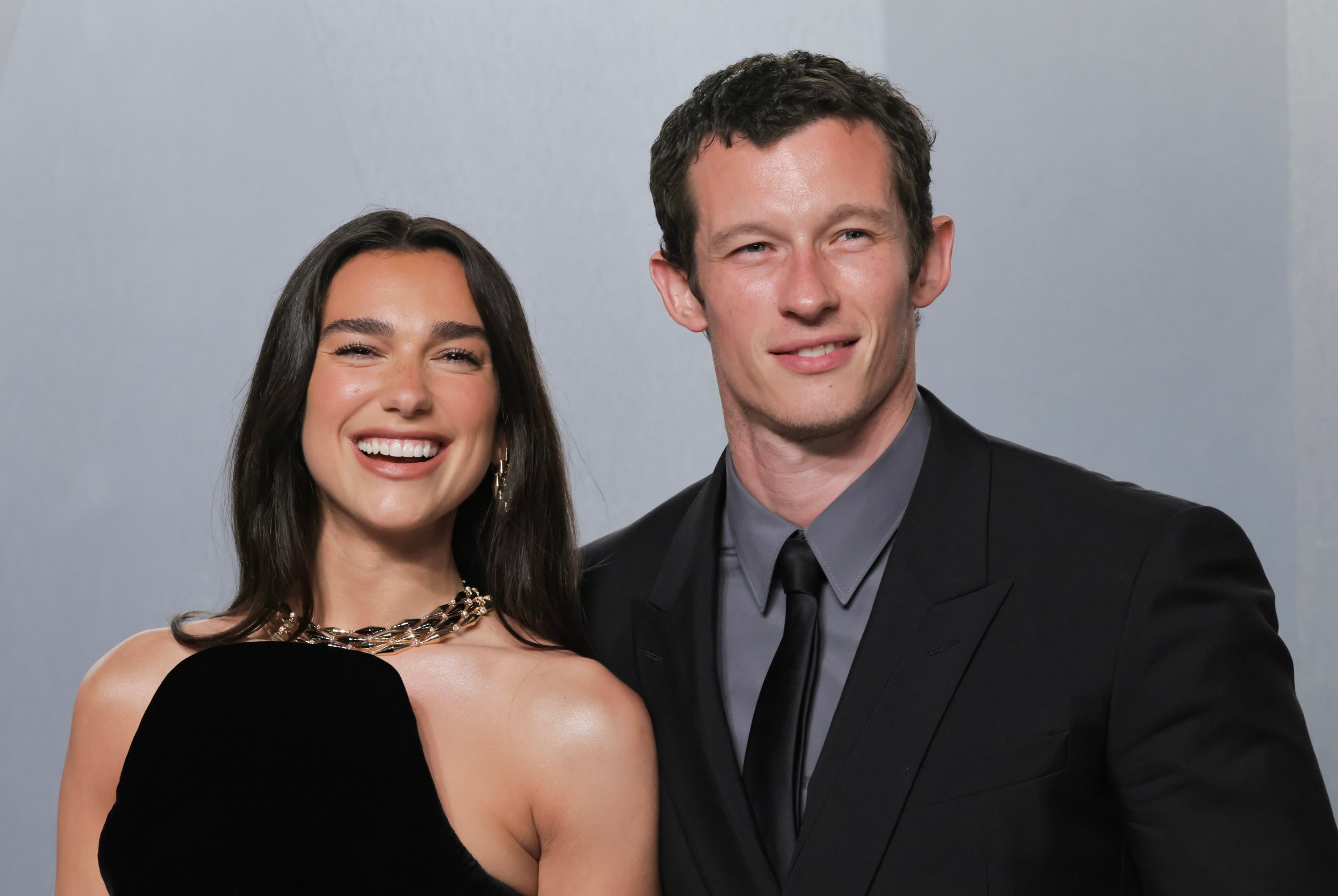 Dua Lipa and Callum Turner smile in front of a grey background. Dua Lipa wears a black dress with a gold chain and Turner wears a black suit with a charcoal grey button-up shirt