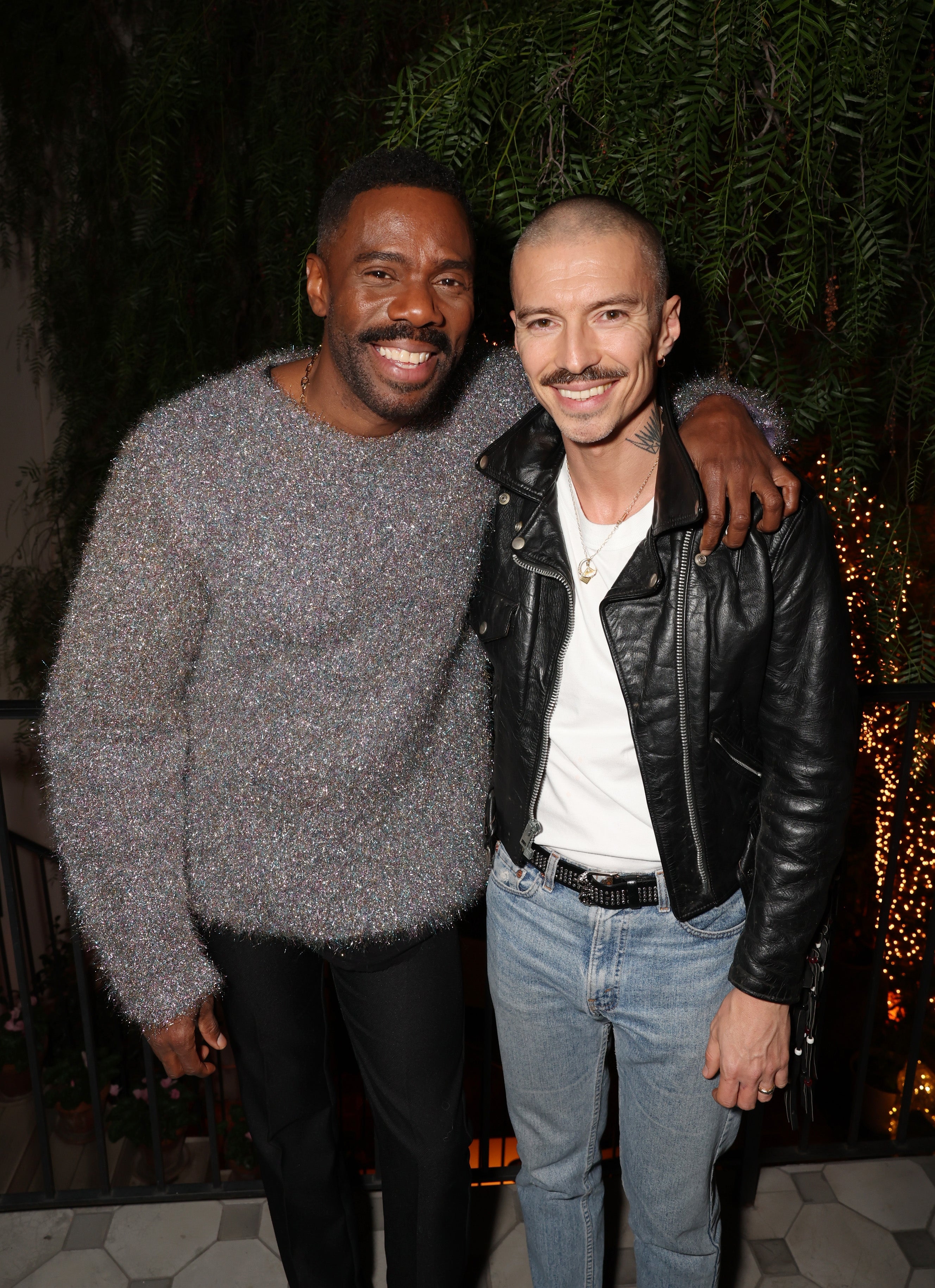 Colman and Raúl Domingo smile as they pose together. Colman is wearing a fluffy sweater, and Raúl is wearing a leather jacket with a white shirt and jeans. They have their arms around each other