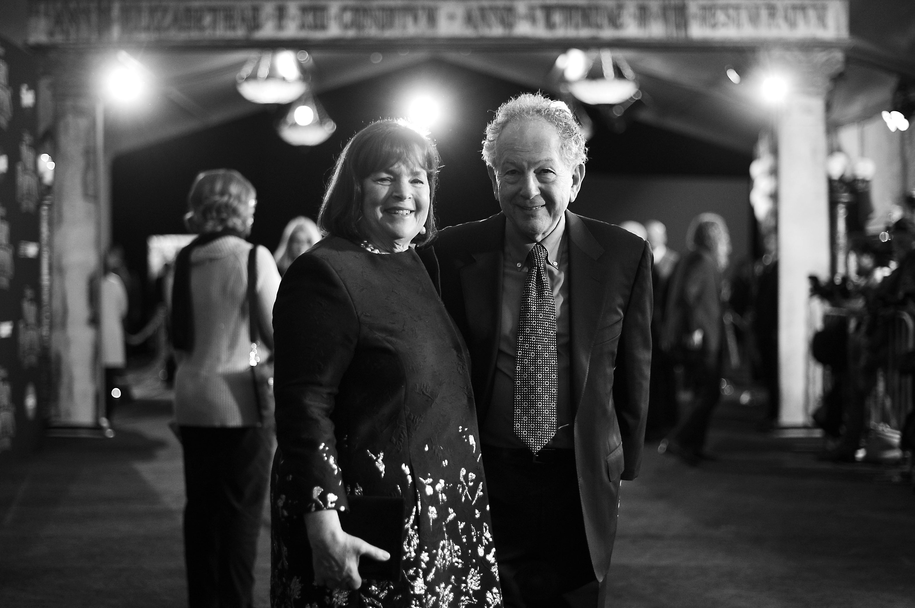 Ina and Jeffrey Garten smile while standing on a red carpet at an event. Ina wears a floral-patterned dress, and Jeffrey wears a suit with a patterned tie