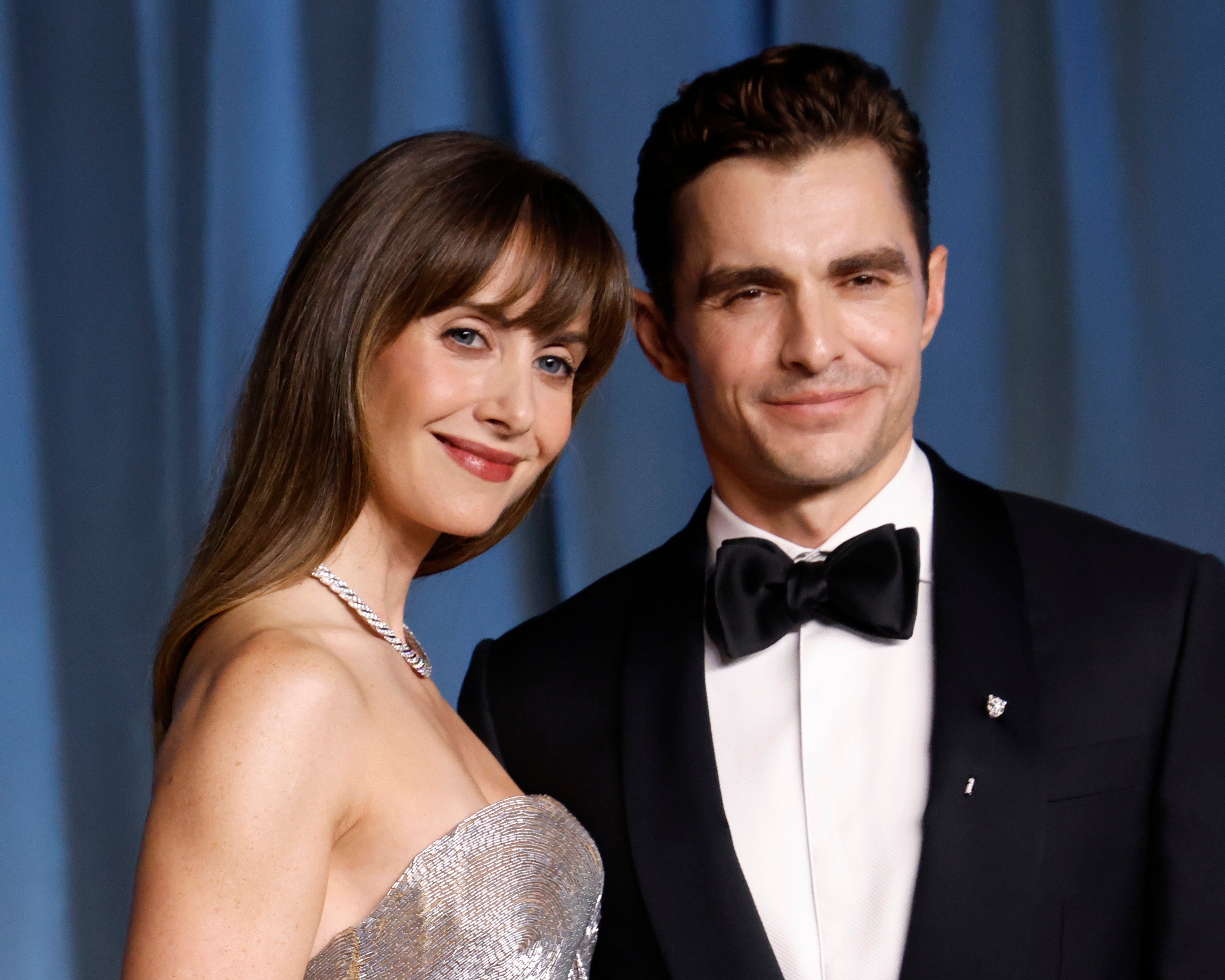 Alison Brie and Dave Franco smile on a formal event carpet. Brie wears an elegant strapless dress, and Franco dons a classic tuxedo with a bow tie