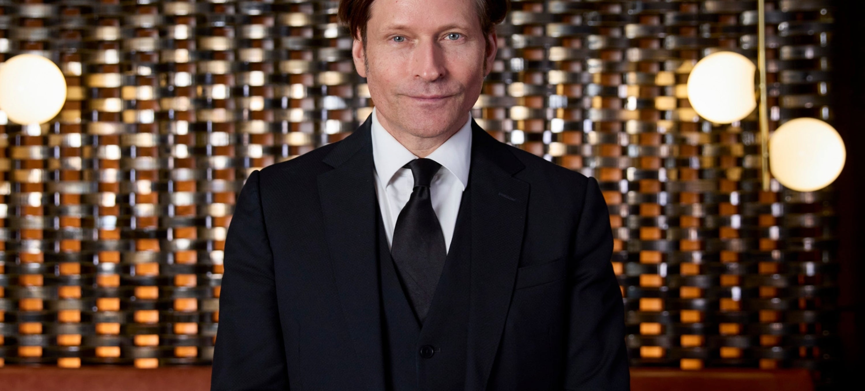 A person in a black suit and tie stands in front of a modern, stylish background with hanging lights