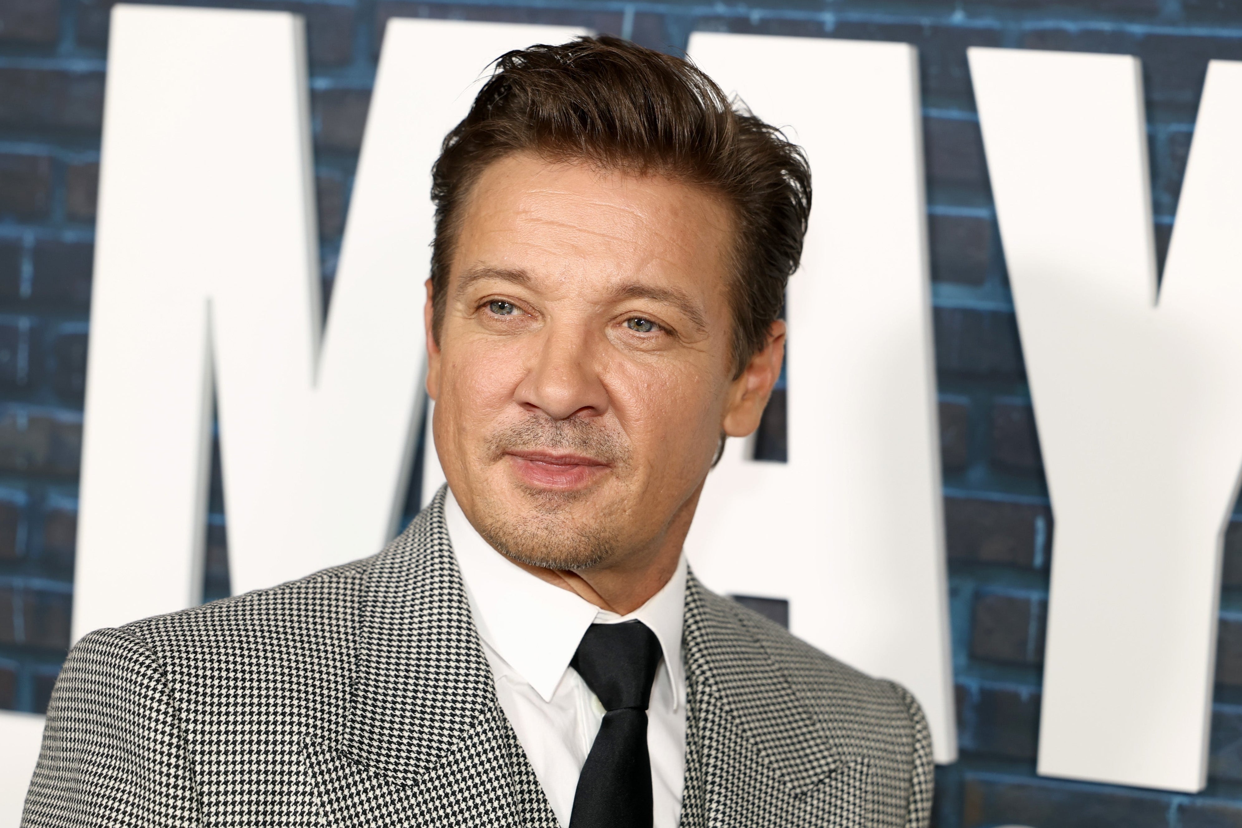 Person in a checkered suit and tie, standing in front of a backdrop with large text, at a celebrity event