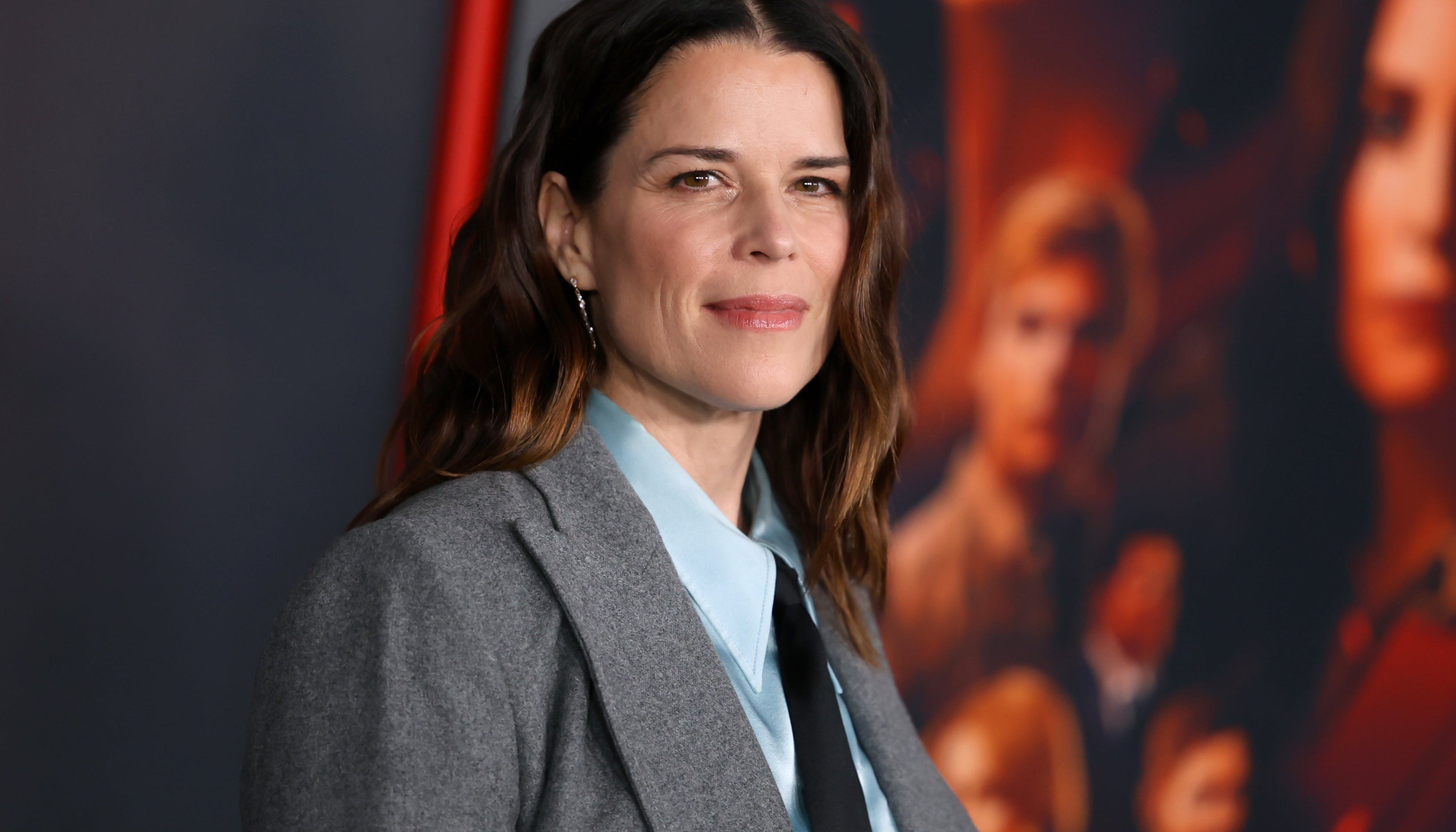A person stands on the red carpet wearing a gray blazer over a blue shirt and black tie, with a backdrop featuring various people