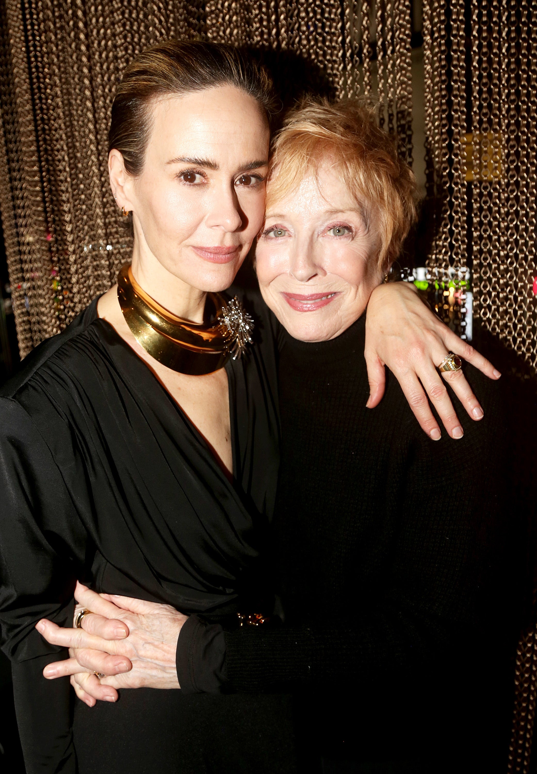 Sarah Paulson and Holland Taylor embrace in front of a gold curtain. Paulson wears a black dress with a bold gold necklace and Taylor wears an elegant black dress
