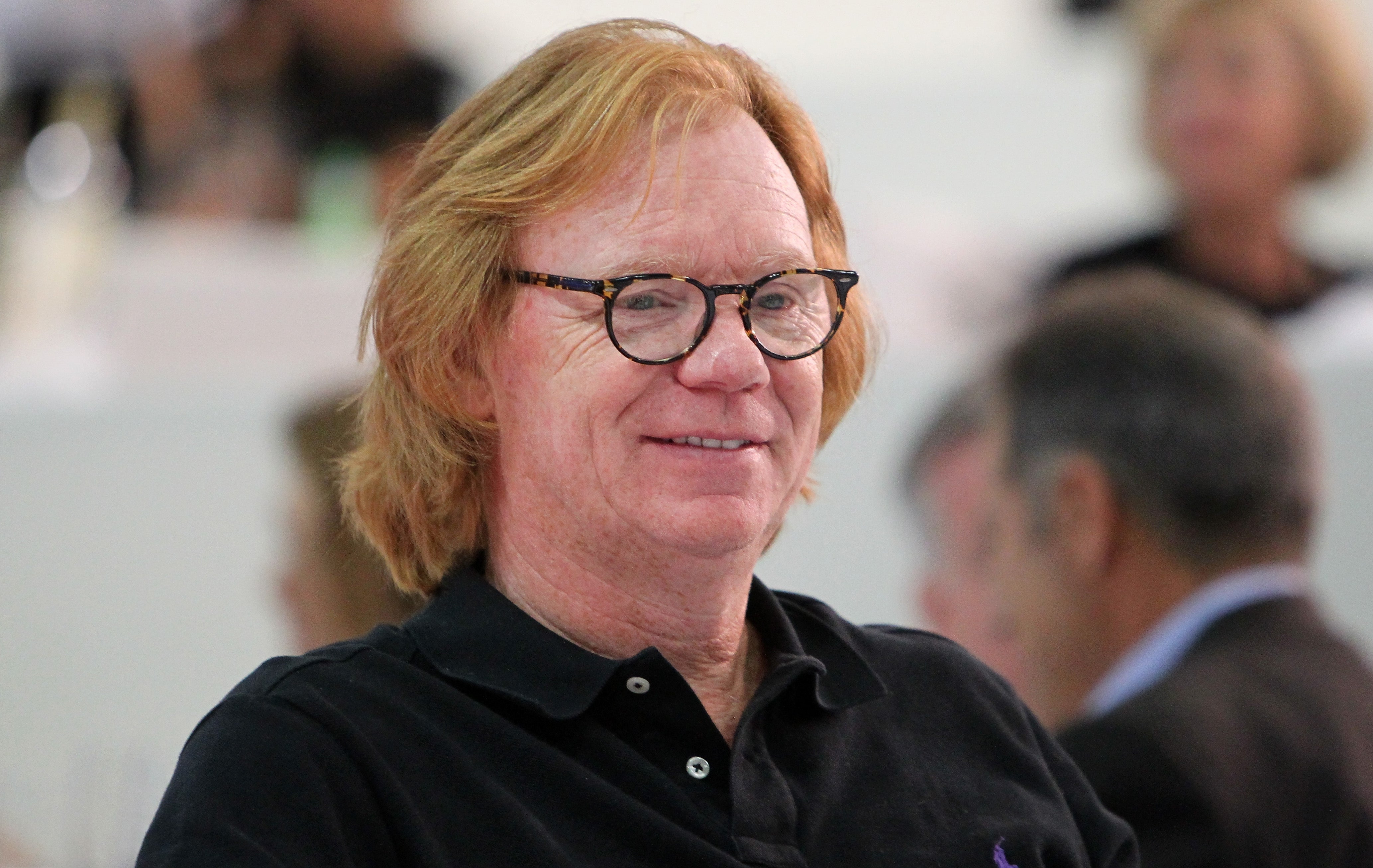 A man with glasses and medium-length hair wears a black polo shirt, smiling at an indoor event