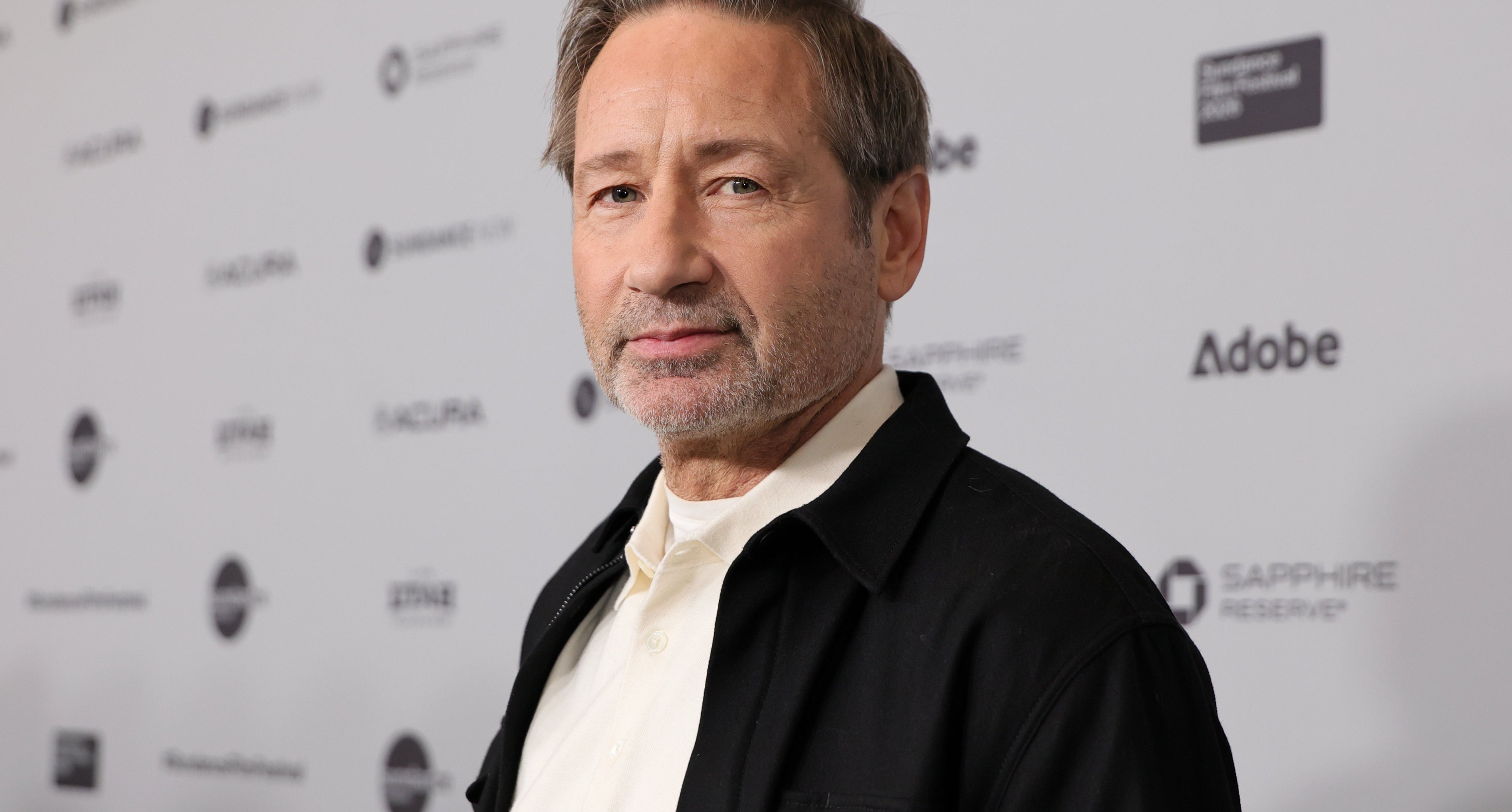 A person in a casual jacket and shirt at a film event, standing in front of a backdrop with sponsor logos