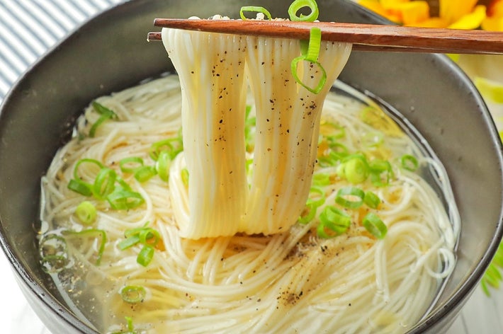 器に盛られた麺を箸で持ち上げる様子。刻んだネギがちらされている。背景にヒマワリが見える。