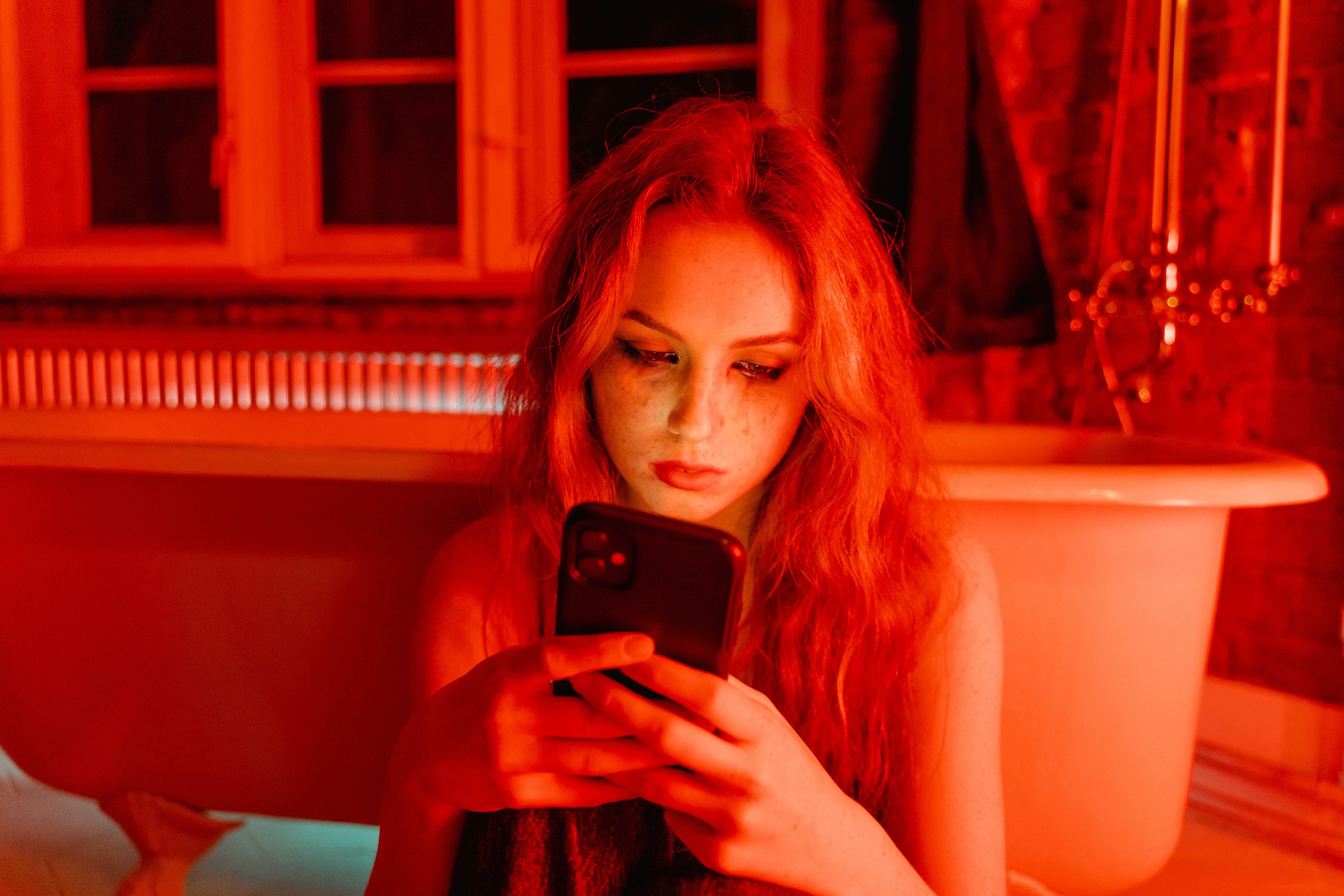 Person med langt hår som leser en telefon på et svakt opplyst bad, nær et vintage badekar, skaper en intim og kontemplativ atmosfære