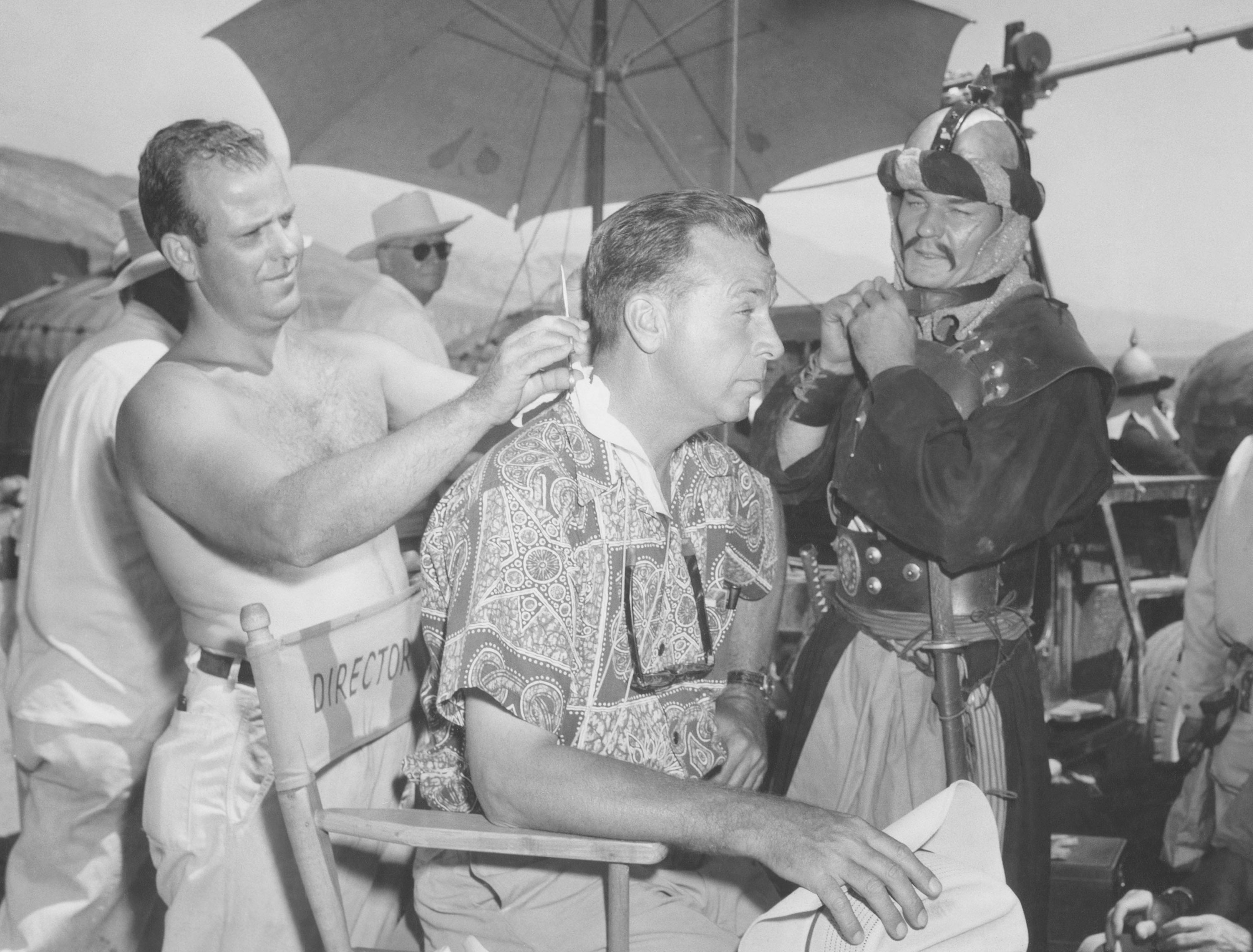 Makeup man Jimmy House cuts Dick Powell's hair on the set of The Conqueror as Leo Gordon watches in culturally insensitive attire