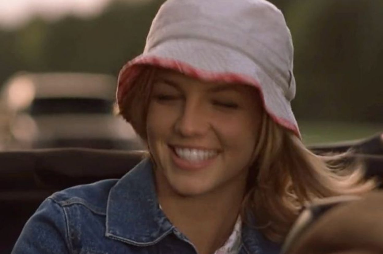 Person smiling in a casual denim jacket and bucket hat while sitting in a moving vehicle outdoors
