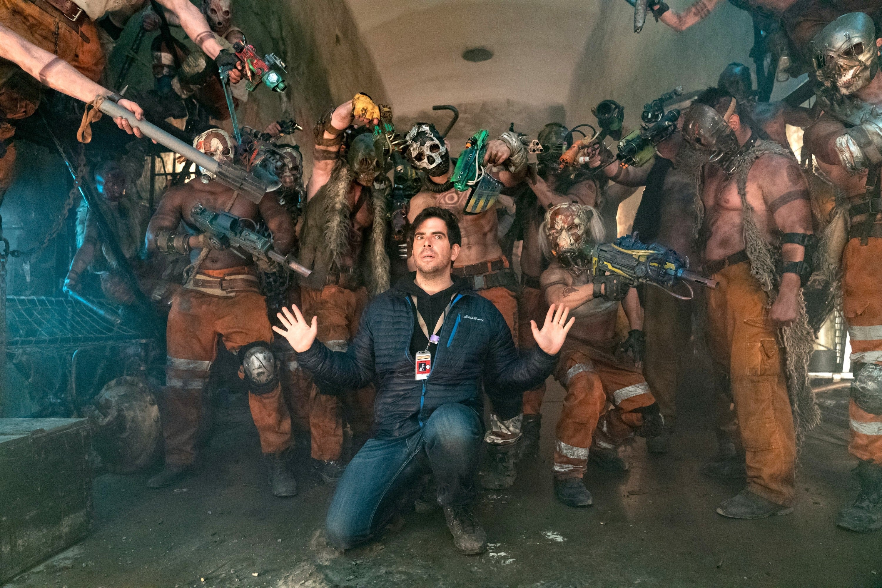 Eli Roth posing in a tunnel surrounded by actors in post-apocalyptic costumes holding weapons at him