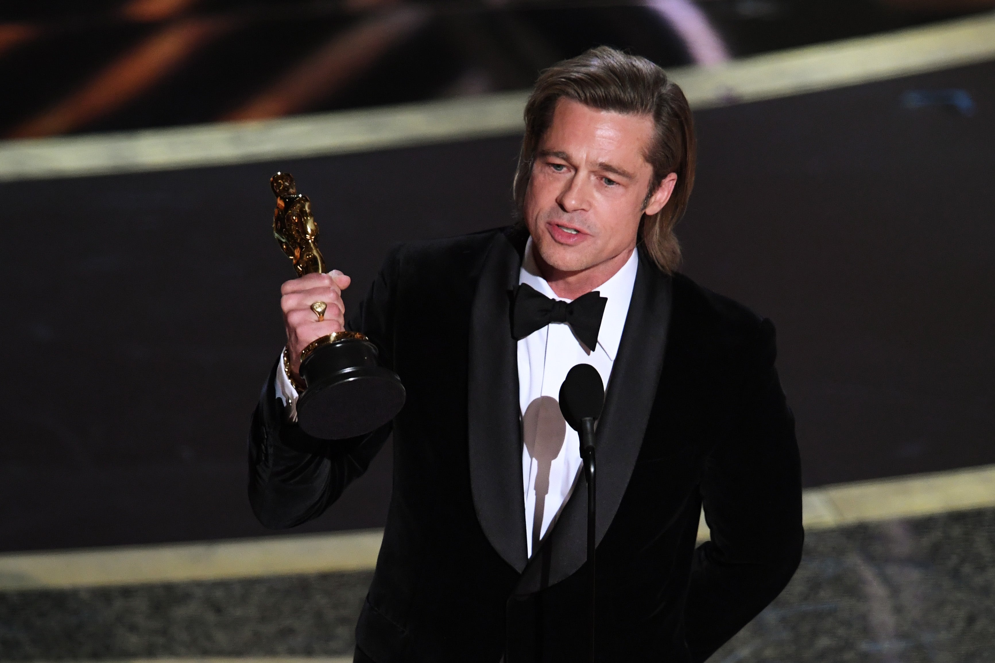 A man in a tuxedo holds an award statue and speaks at a microphone on stage