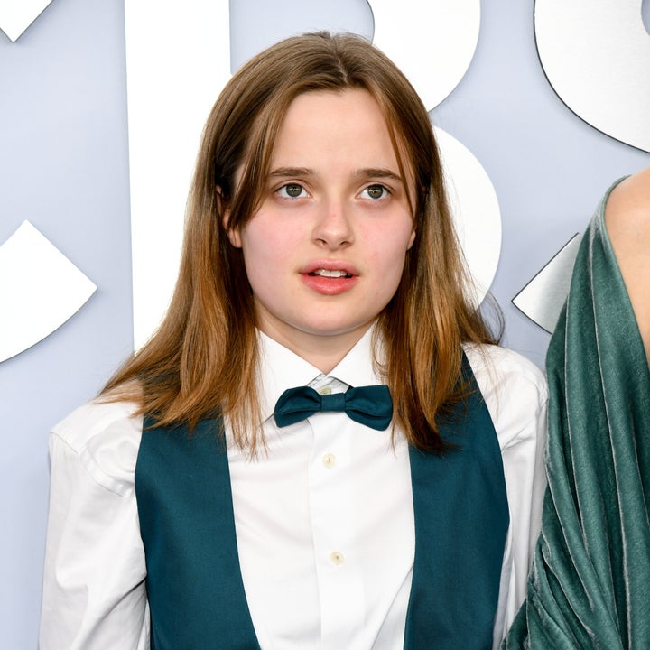 Person on a red carpet wearing a white shirt with a bowtie and vest. They have long hair and a neutral expression. Velvet fabric visible nearby