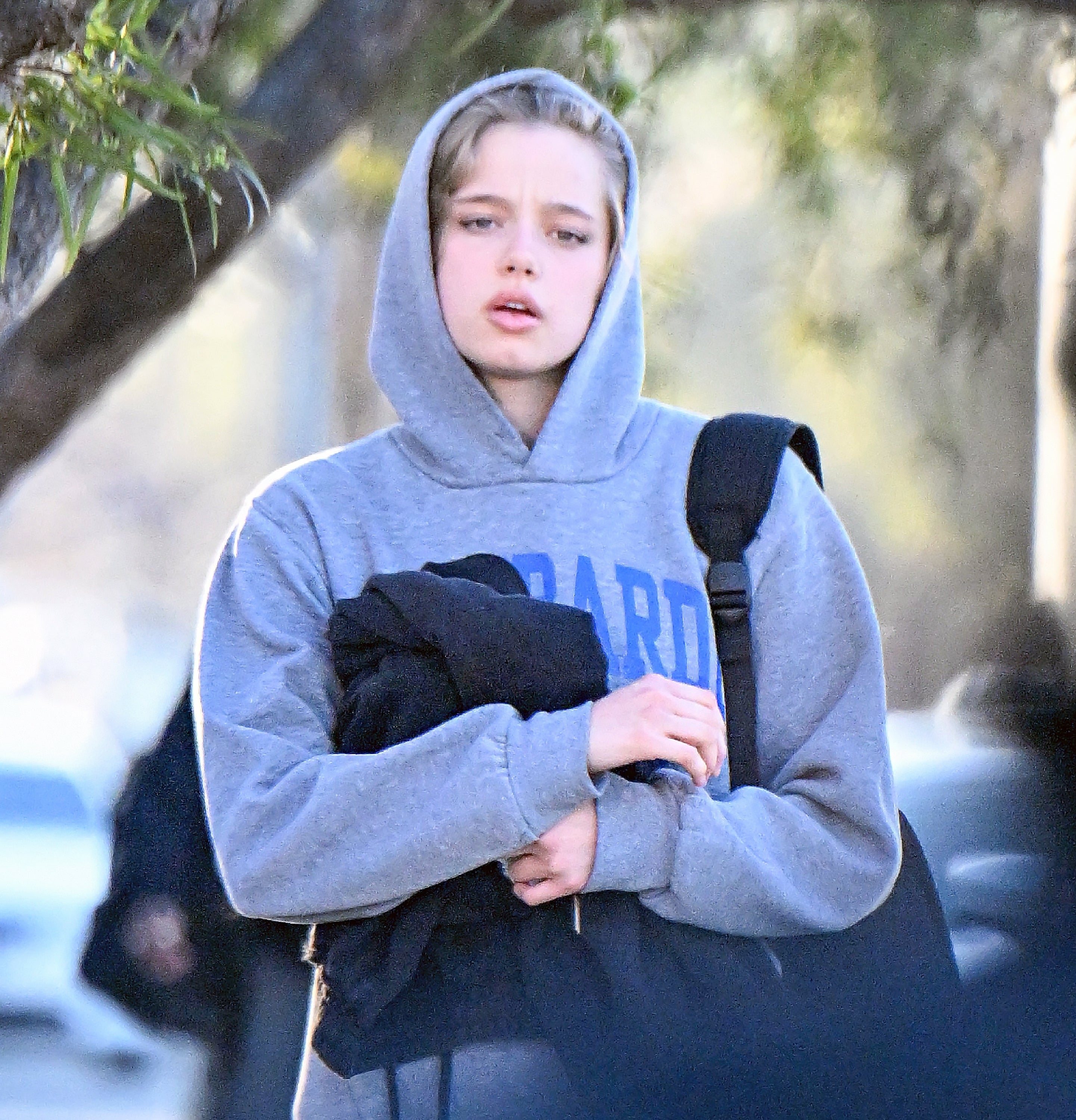 Person walking outdoors, wearing a hooded sweatshirt, holding a jacket and a bag. Trees are in the background