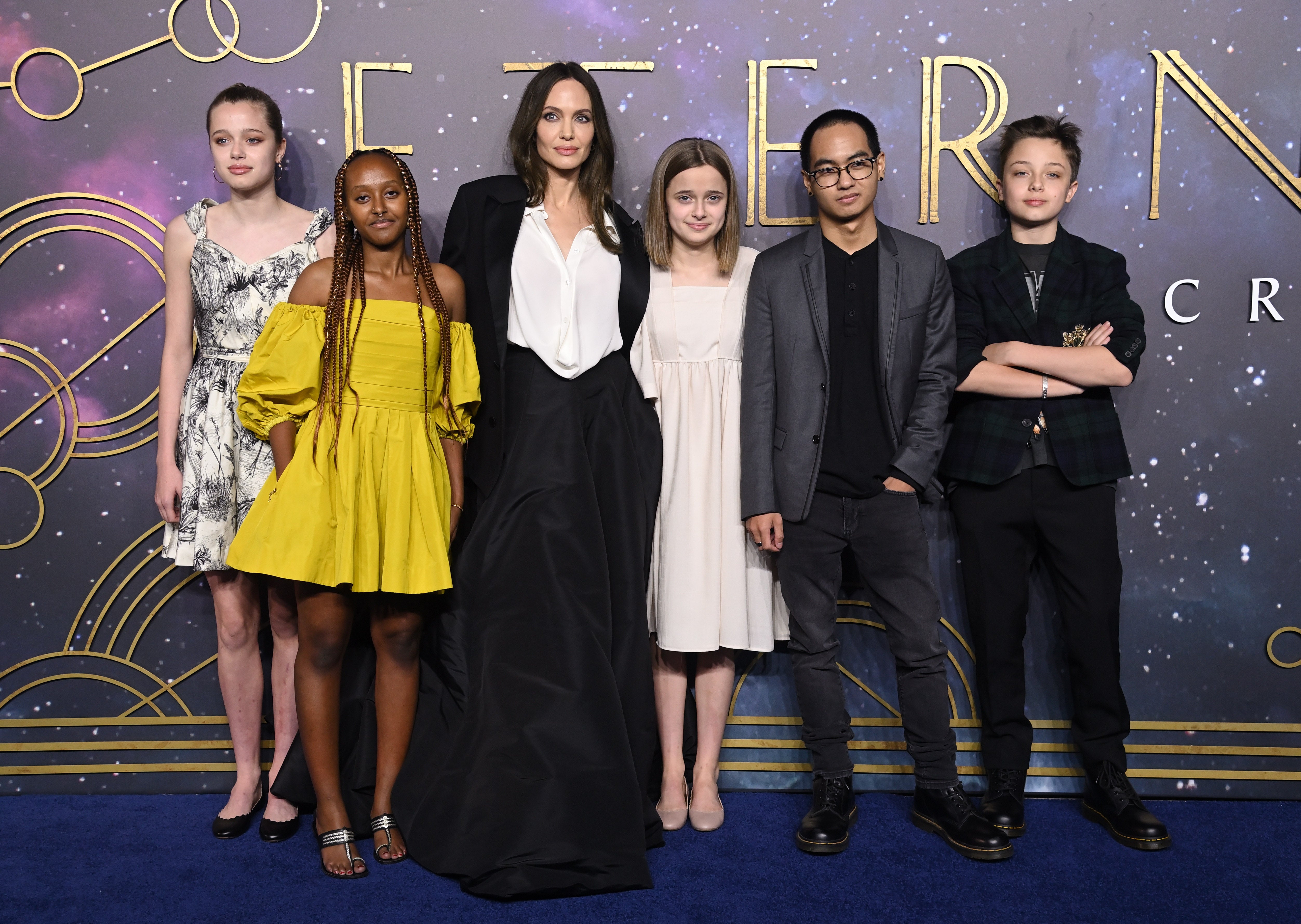 A group of six people stands on a red carpet. One is wearing a white shirt with a long black coat. Others are in various outfits, including a yellow dress