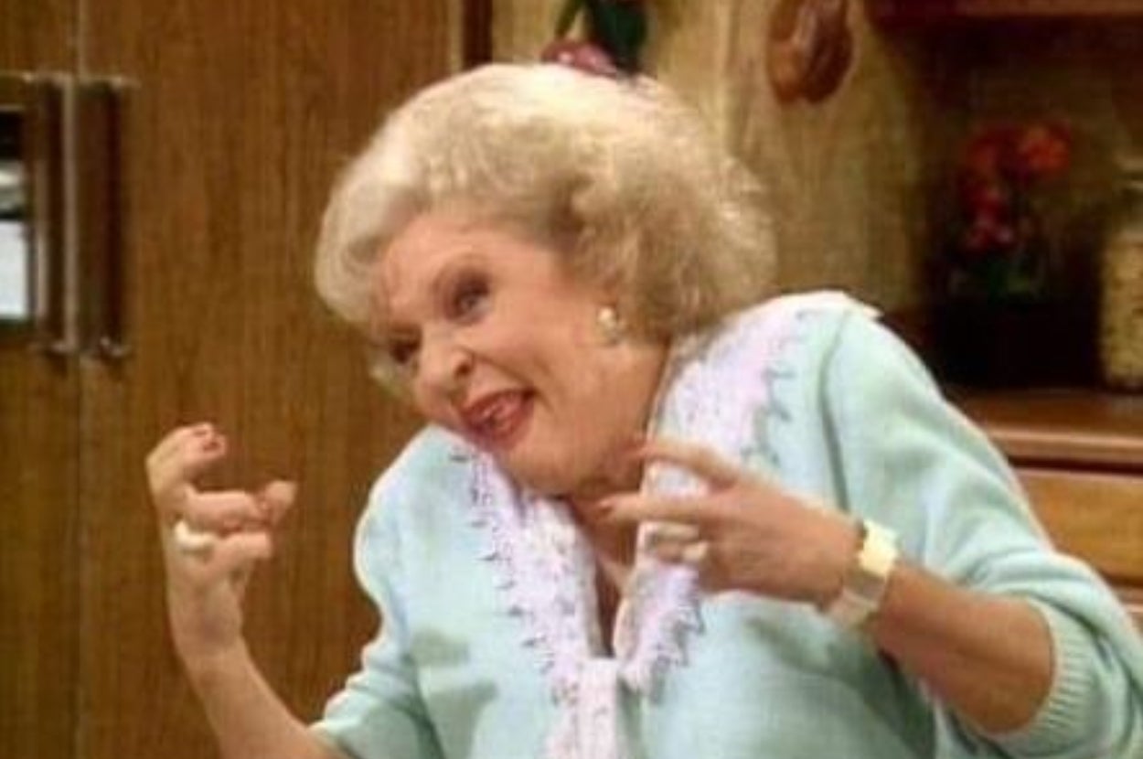 Elderly woman with short curly hair, wearing a light-colored blouse, makes an animated expression with her hands raised, inside a home setting