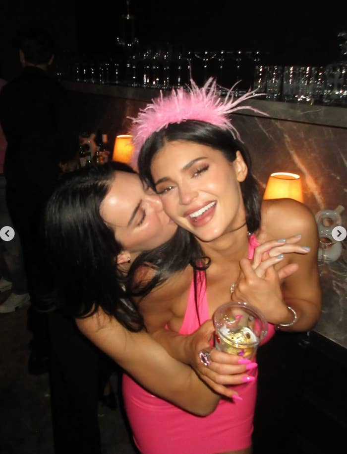 Two women smiling and embracing at a party, one wearing a playful feathered headband and the other giving a cheek kiss. They appear to be enjoying themselves