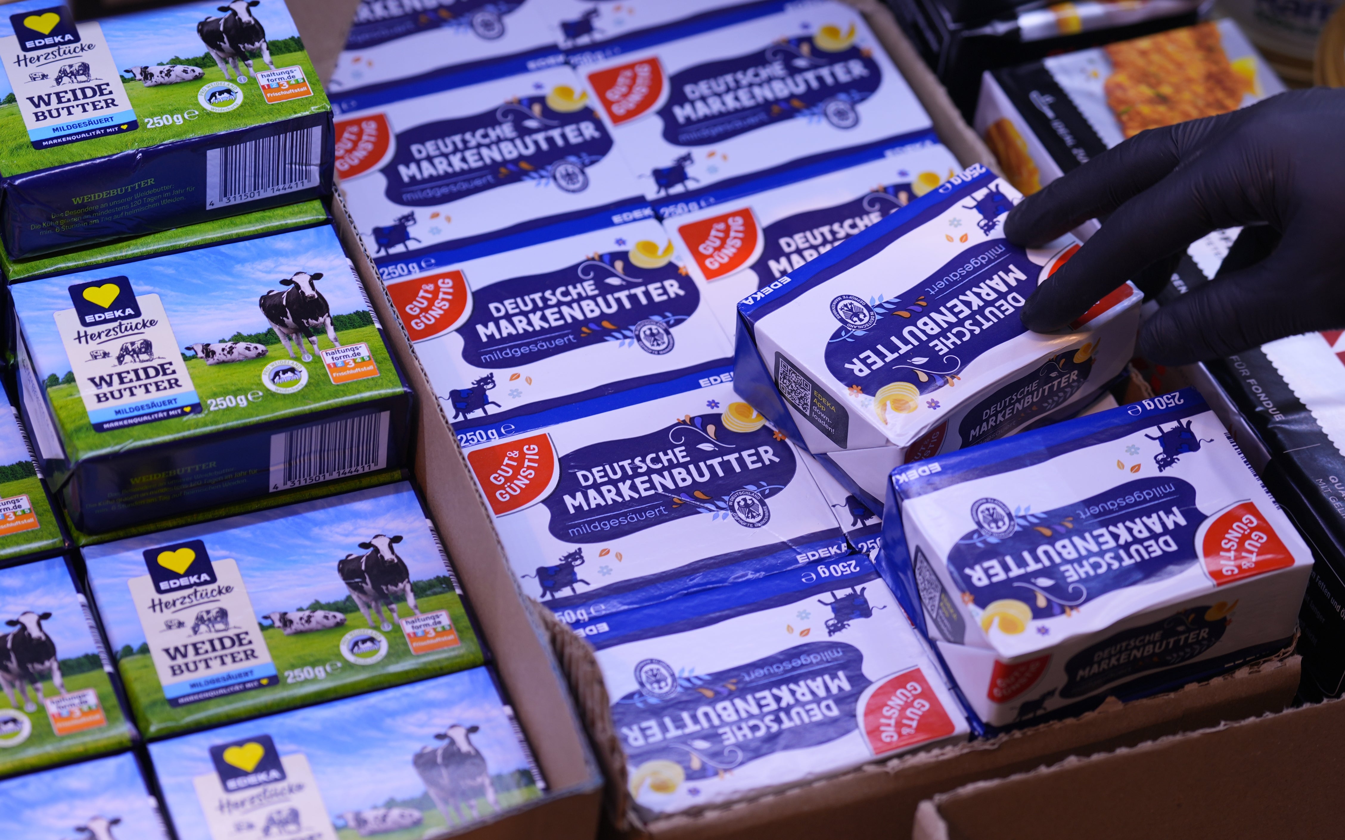 Esker med Deutsche Markenbutter stablet i en butikk med en hånd som strekker seg etter en. I nærheten ligger Herzstücke smørpakker med en kuillustrasjon