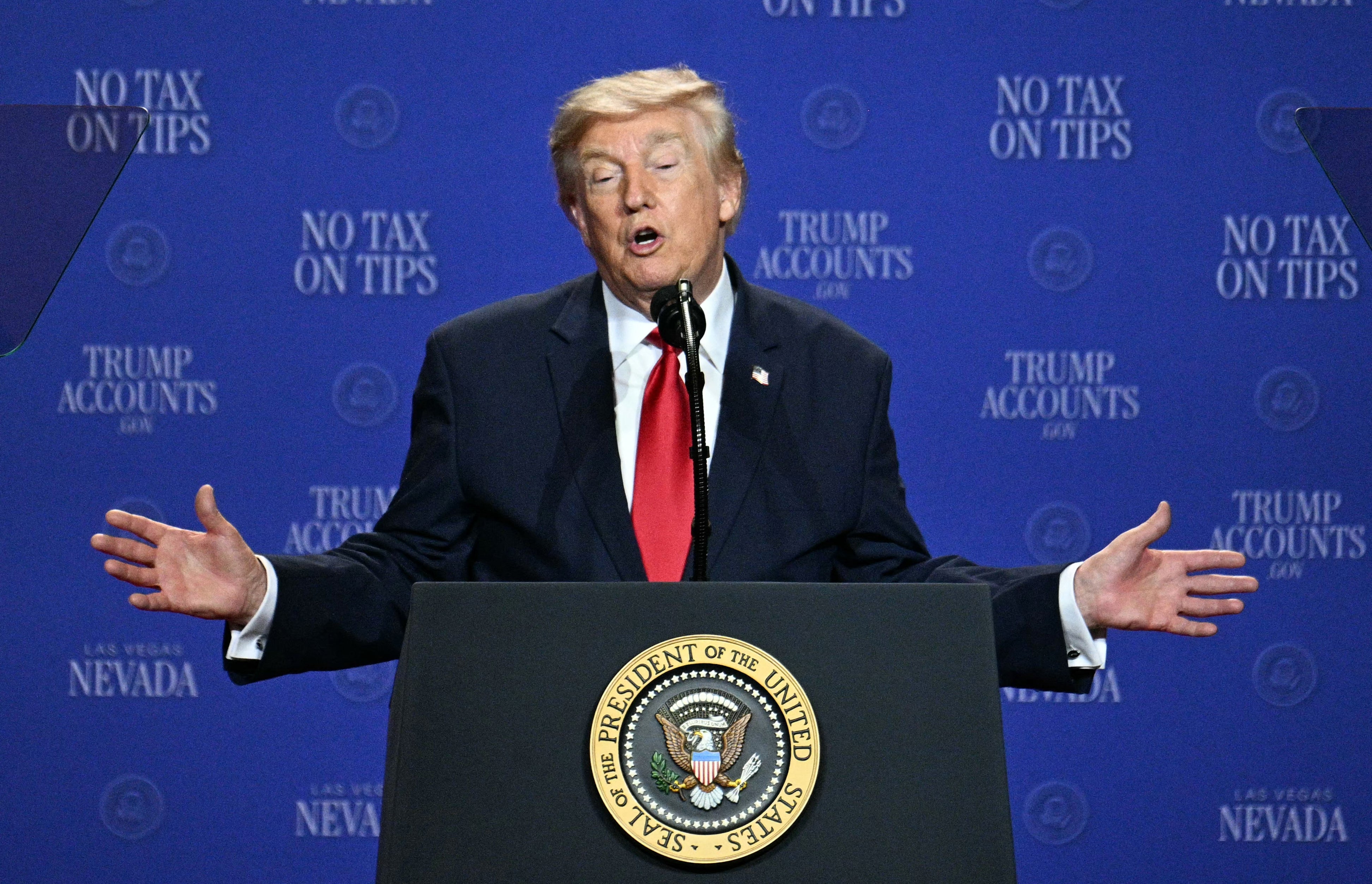 En person i dress og rødt slips snakker ved et podium med «Ingen skatt på tips»; og "Trump-kontoer" bannere i bakgrunnen