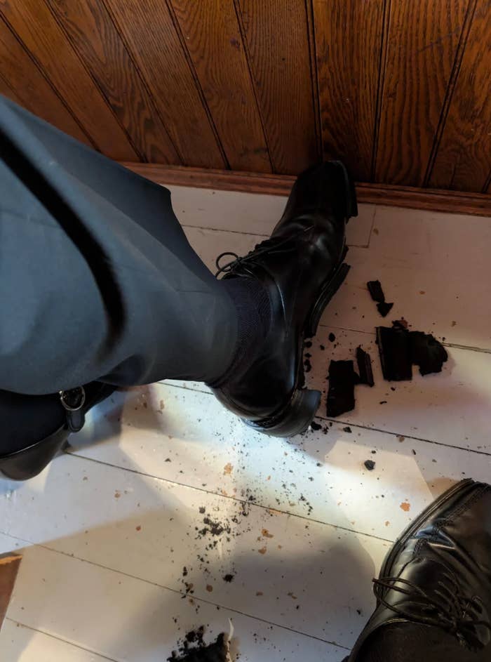 Shoe crushing through floorboards, leaving debris around. Visible part of a suited leg