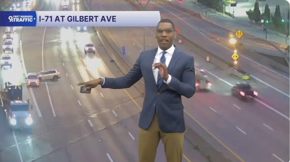 Nyhetsoppleser rapporterer direkte trafikk med biler på I-71 og Gilbert Ave. Iført blazer og bukser, gestikulerer til trafikkflyten på veier