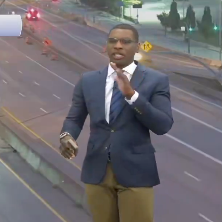 A person stands on a highway appearing to report news, wearing a suit jacket, tie, and holding a phone. Traffic cones are visible in the background