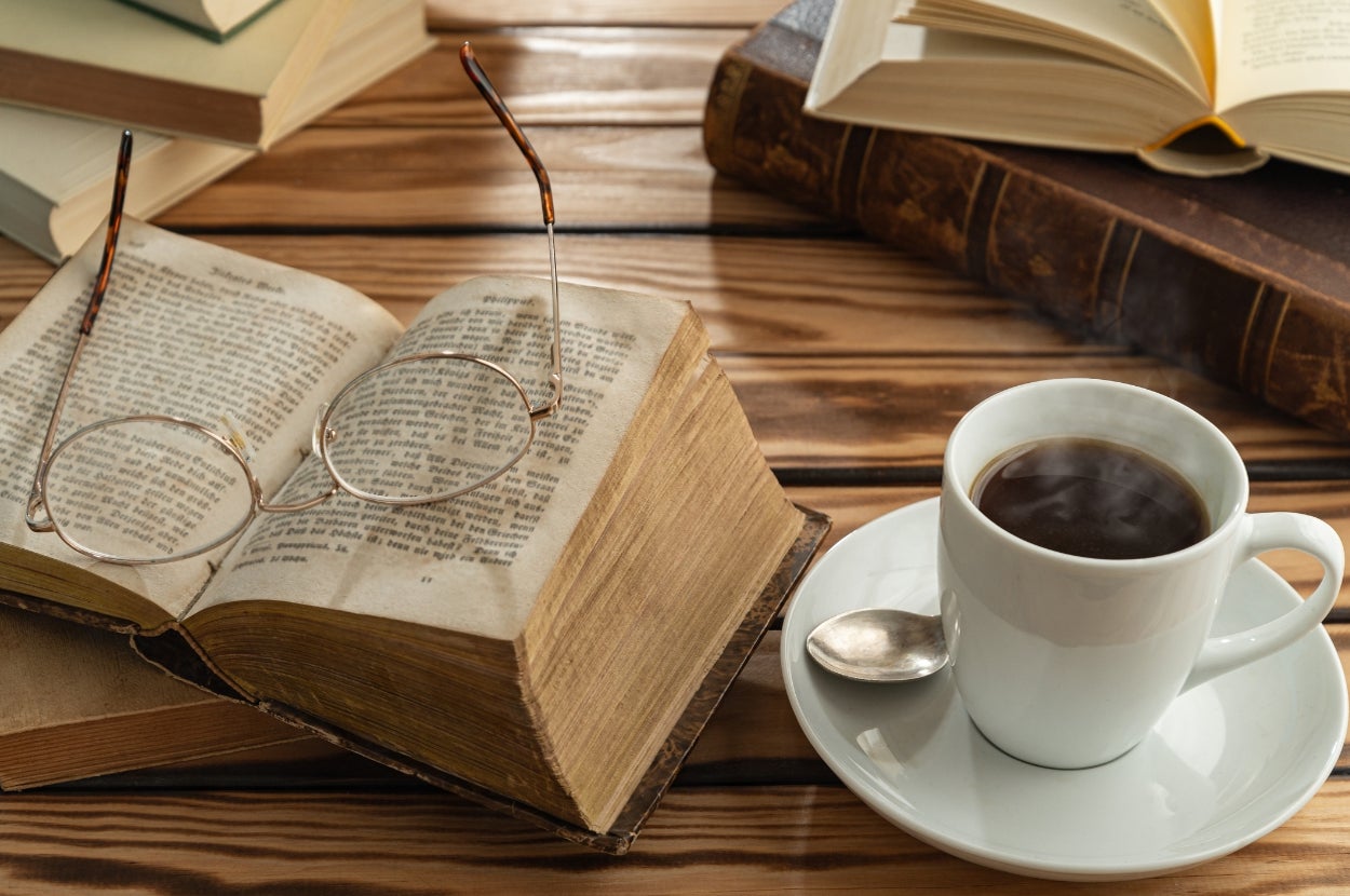 Open book with reading glasses on top, surrounded by other books on a wooden table. A steaming cup of coffee sits on a saucer beside them