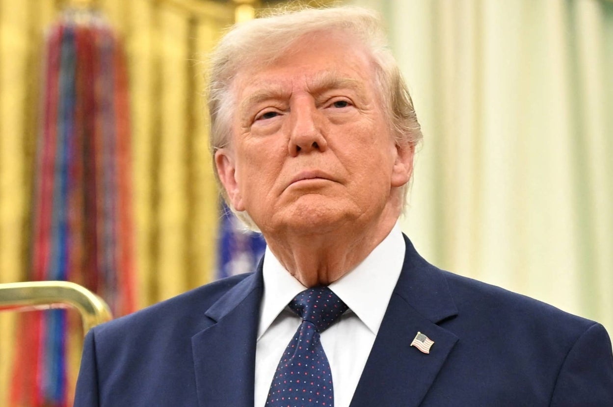 A person in a suit and tie stands with a serious expression indoors