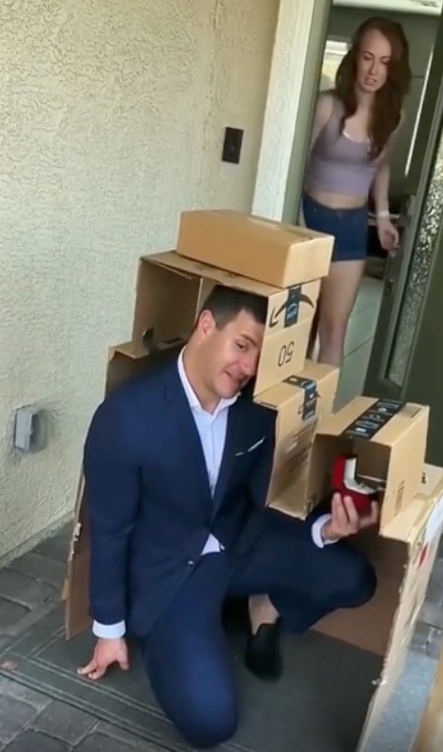 A person in a suit kneels, holding a ring box under a stack of cardboard boxes on a doorstep, as another person looks on from the doorway