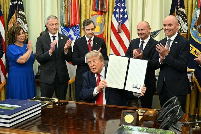 Donald Trump at a desk holding a signed document while others stand nearby clapping in a room with flags and official emblems
