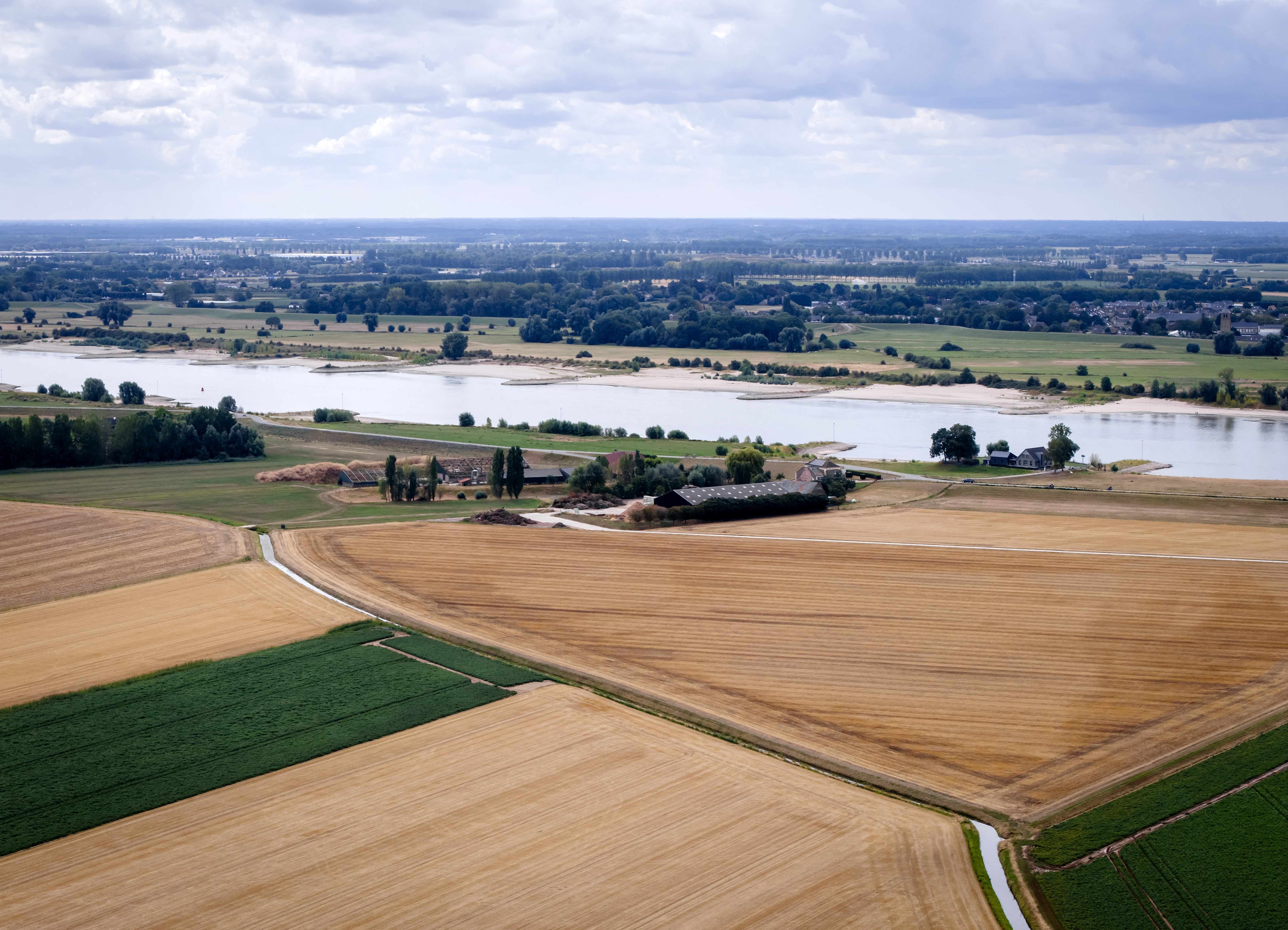 Luftfoto av vidstrakte jordbruksland med en elv som renner gjennom, oversådd med trær og noen få bygninger. Overskyet himmel