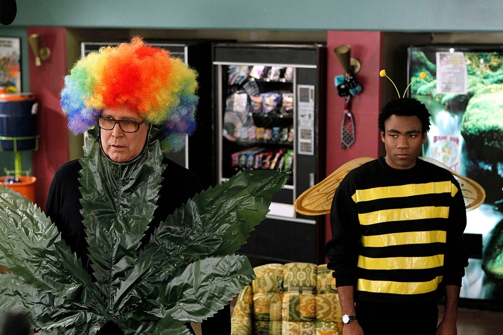 Chevy Chase and Donald Glover in a colorful costume scene: Chevy dressed as a leafy plant with a rainbow wig, the Donald as a bee in a striped top and wings