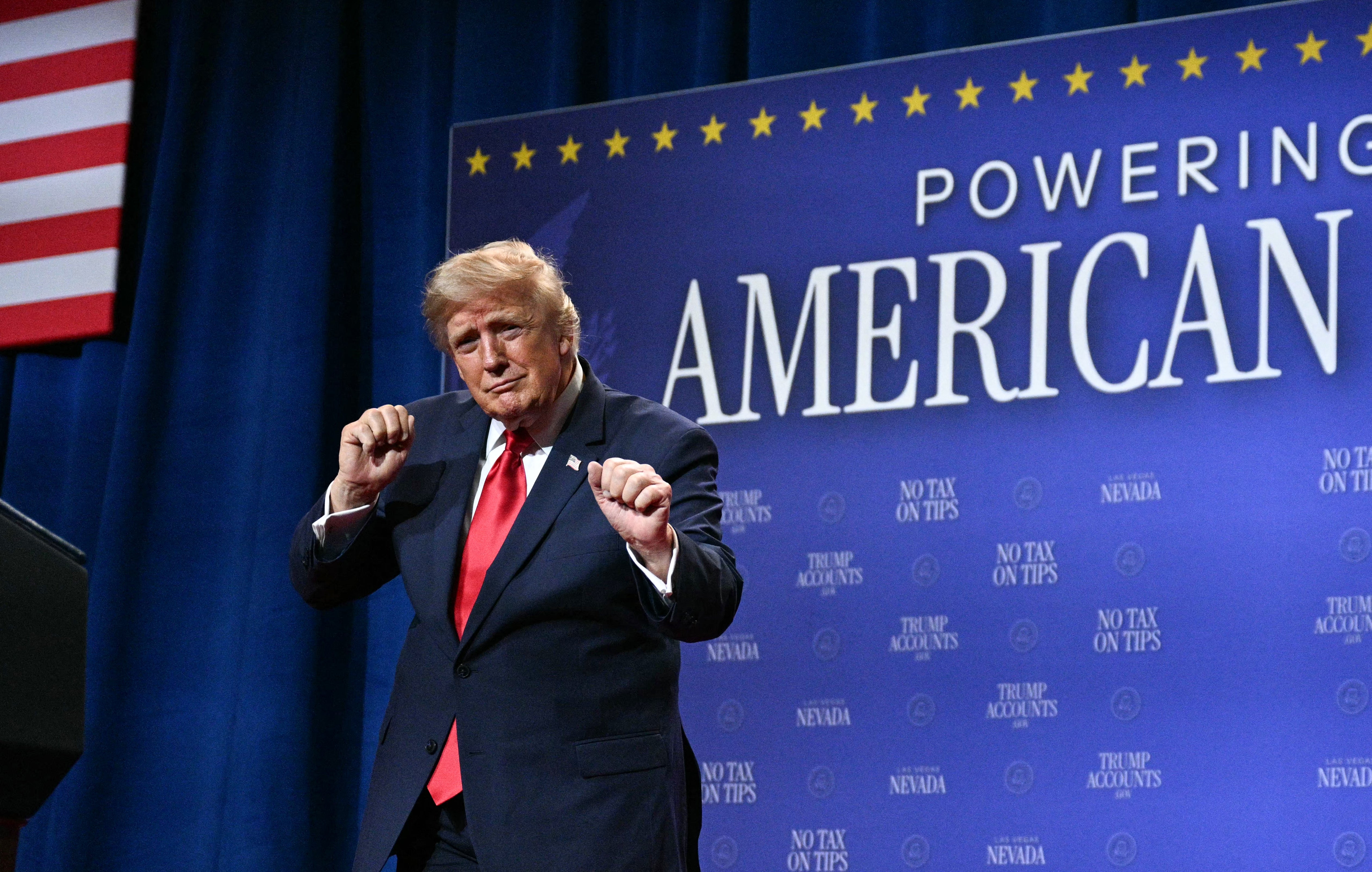 A man in a suit on stage gestures with his fists raised. A background displays phrases like "Powering American" and "No Tax on Tips."
