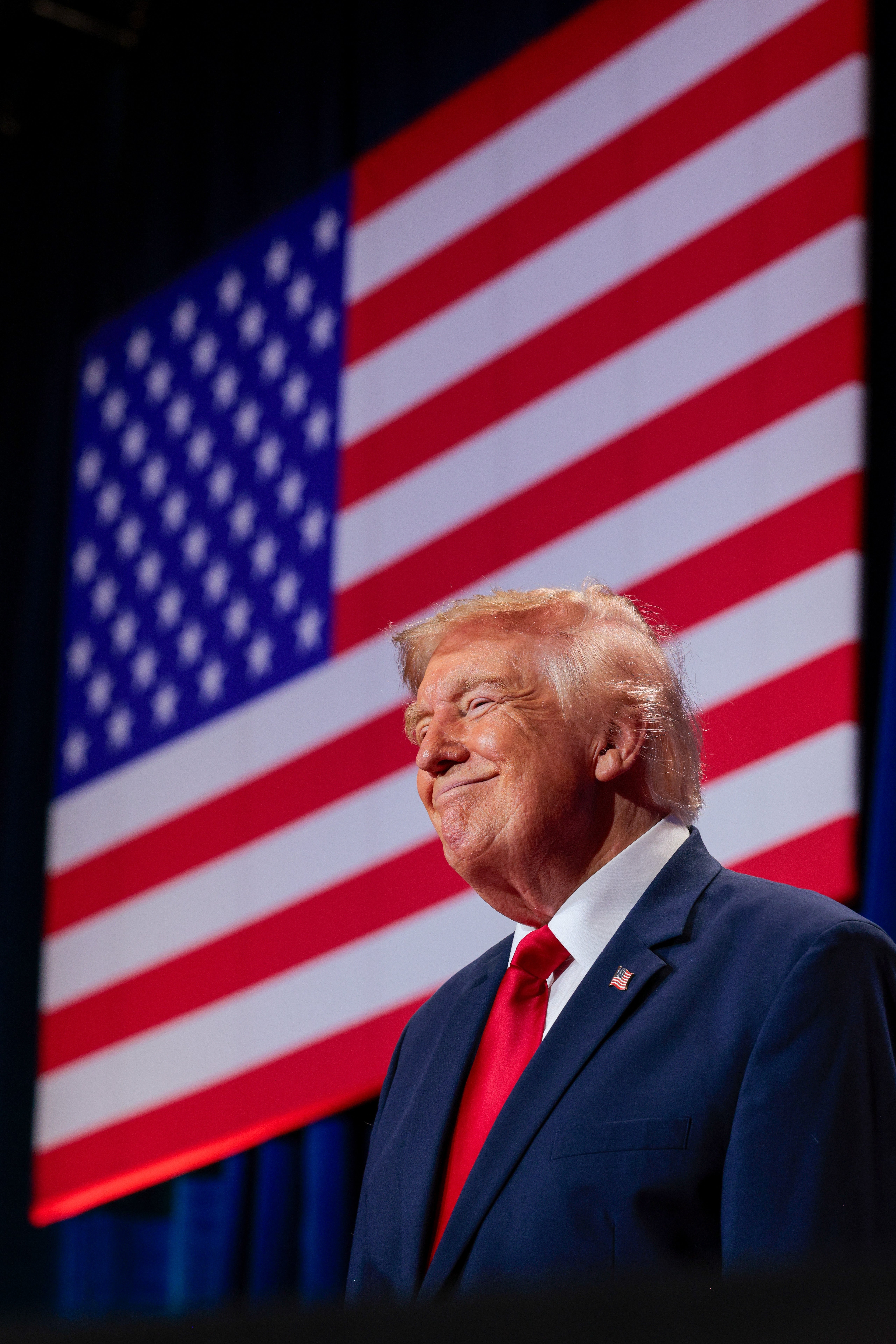 En mann i dress og slips står og smiler foran et stort amerikansk flagg i bakgrunnen