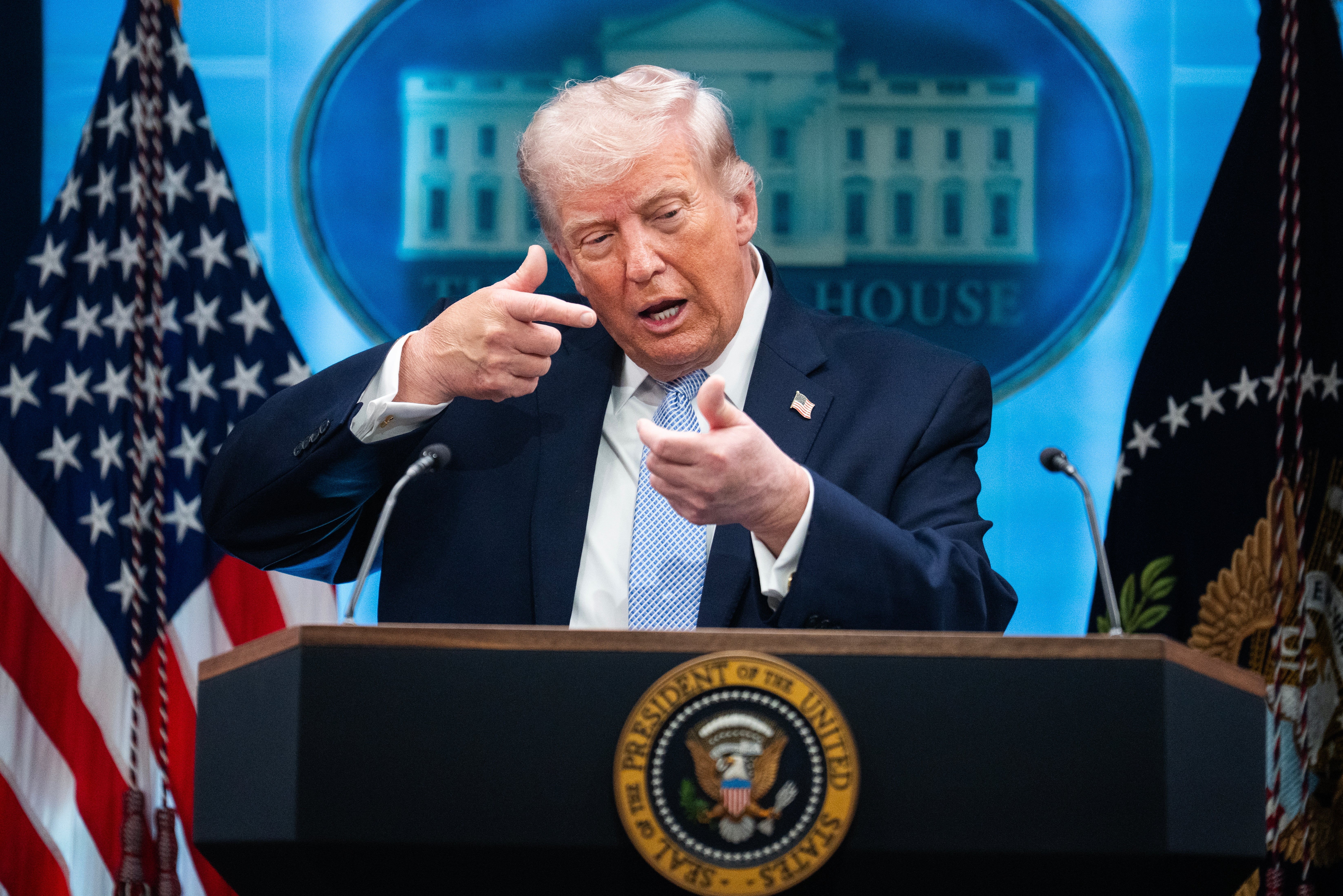 En person på et podium i dress gestikulerer med hendene under en pressebriefing, flankert av amerikanske flagg og med Det hvite hus-emblem i bakgrunnen