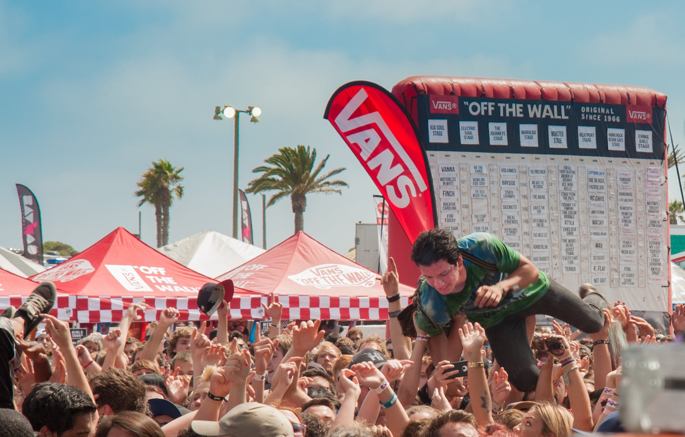 An audience at the Vans Warped Tour, tents and an inflatable with set times is in the background