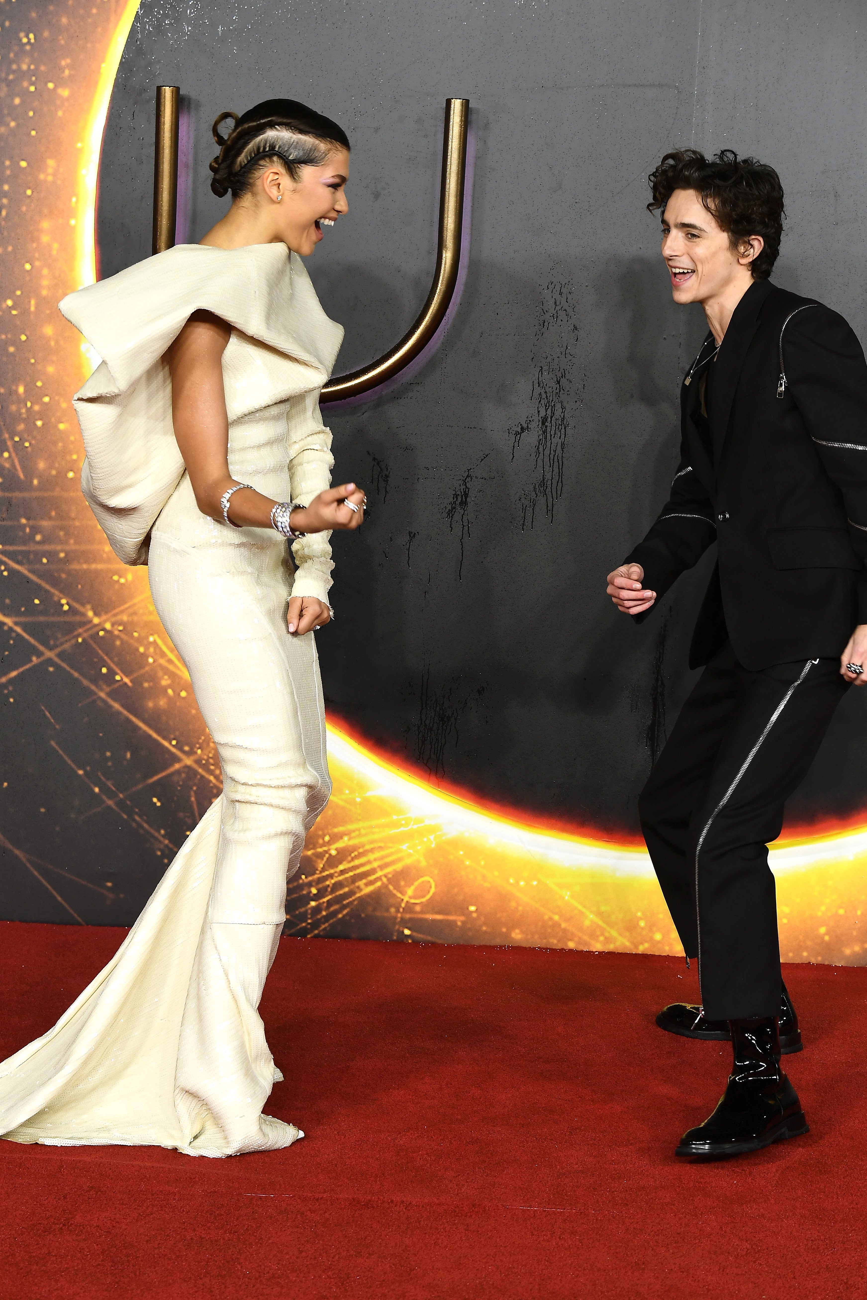 Two individuals on a red carpet, one in an elegant gown with bold shoulders, the other in a stylish suit with zip details, both smiling energetically