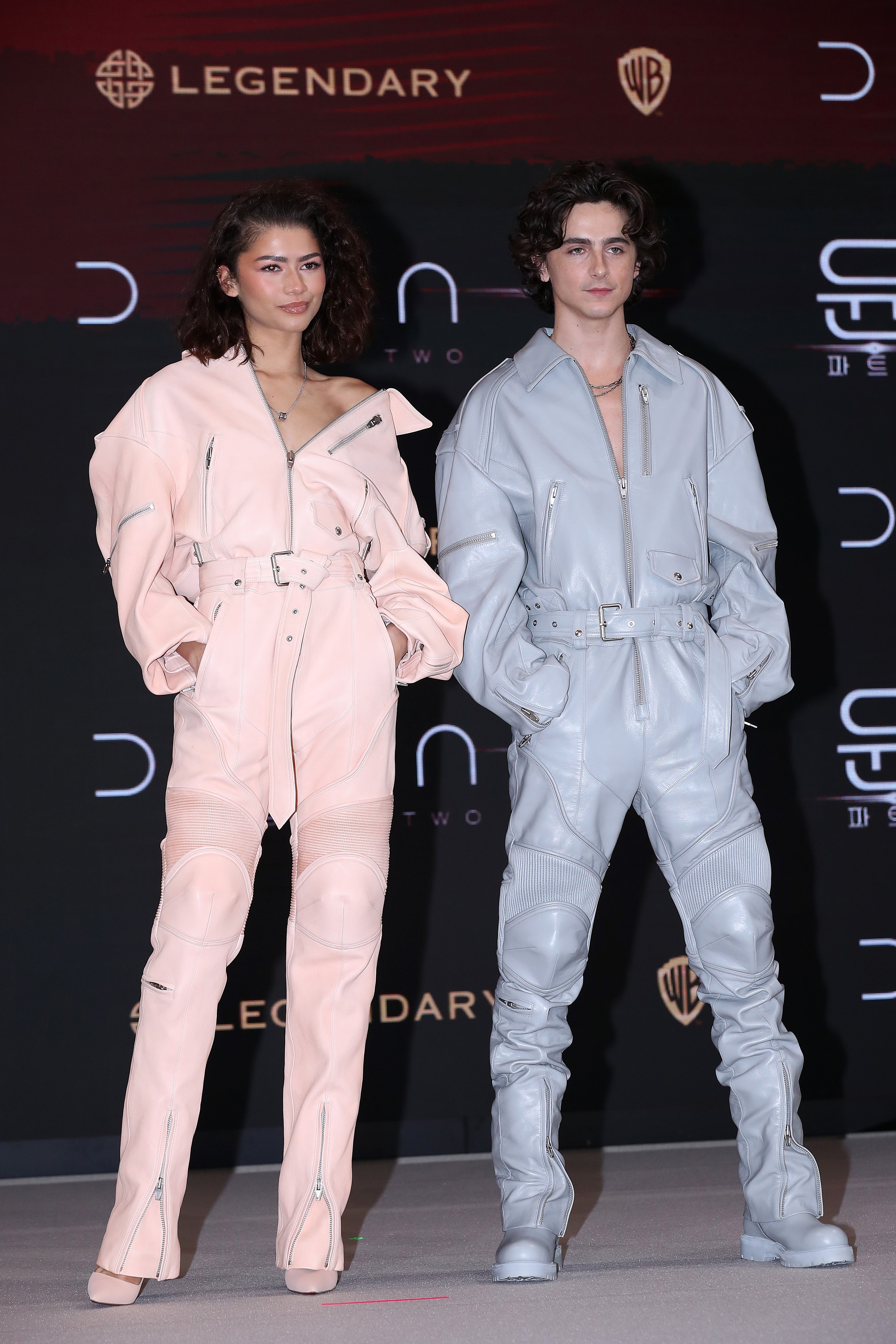 Two individuals on a red carpet wearing stylish, futuristic jumpsuits with structured designs