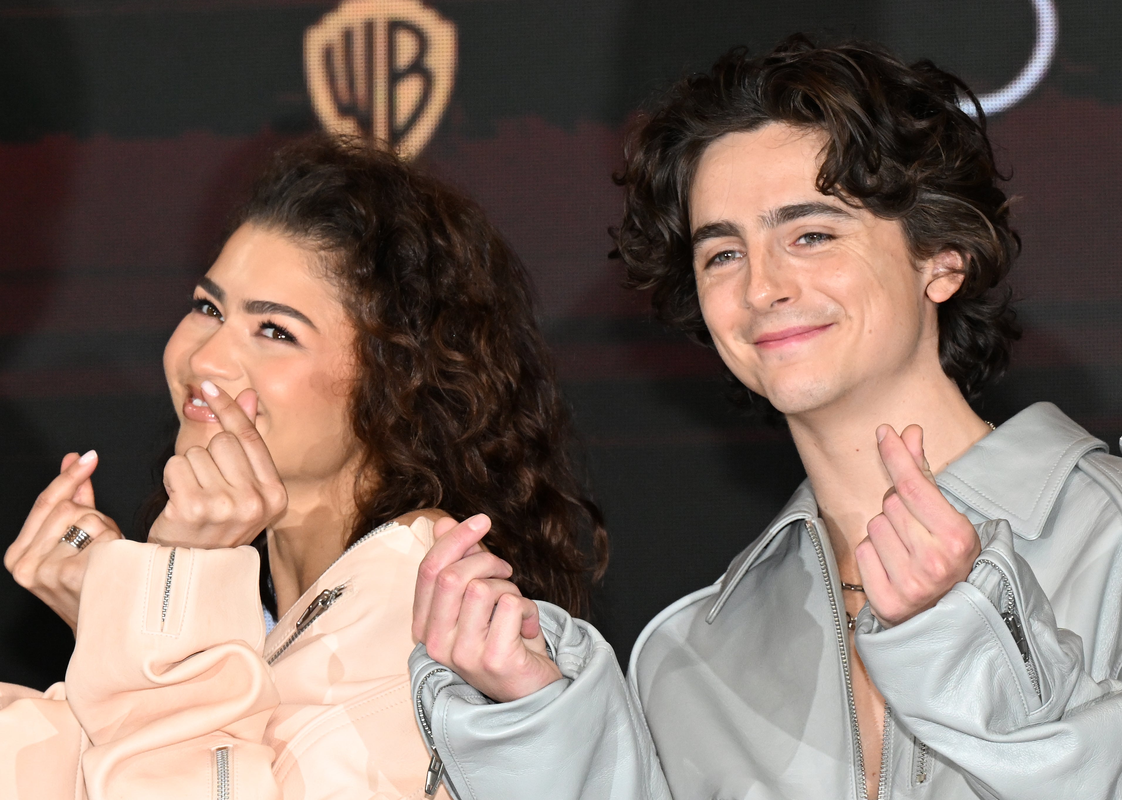 Two individuals on a red carpet making heart gestures with their fingers, both wearing stylish jackets