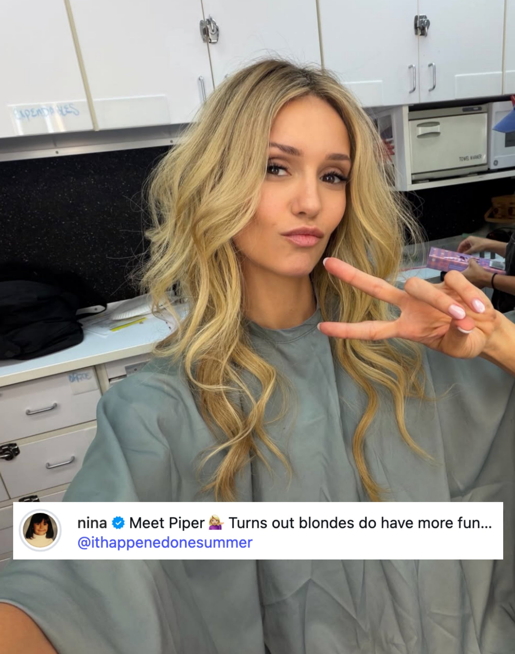 Person in a makeup room, wearing a salon cape, making a peace sign. They have wavy hair and appear relaxed