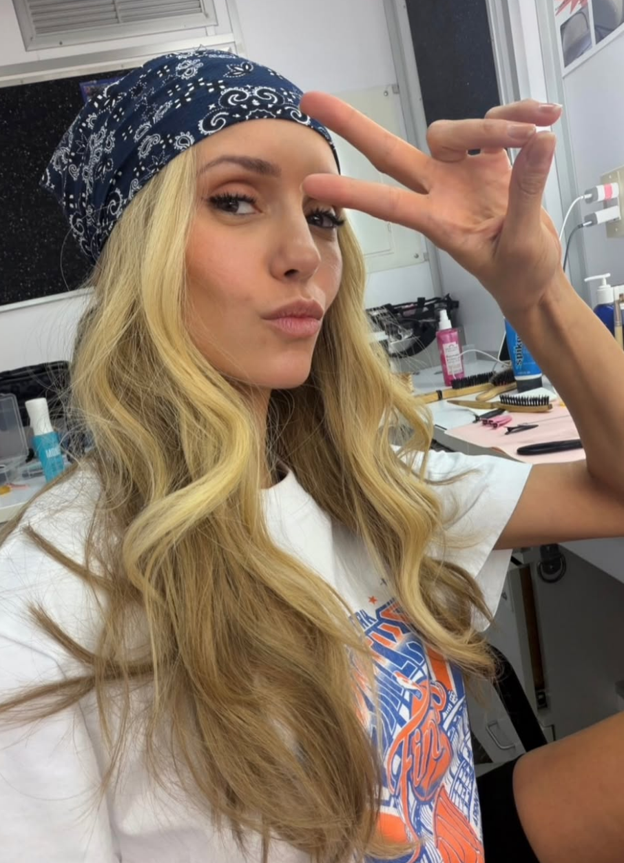 Person with long, wavy hair wearing a bandana and a casual T-shirt poses with a peace sign in a dressing room