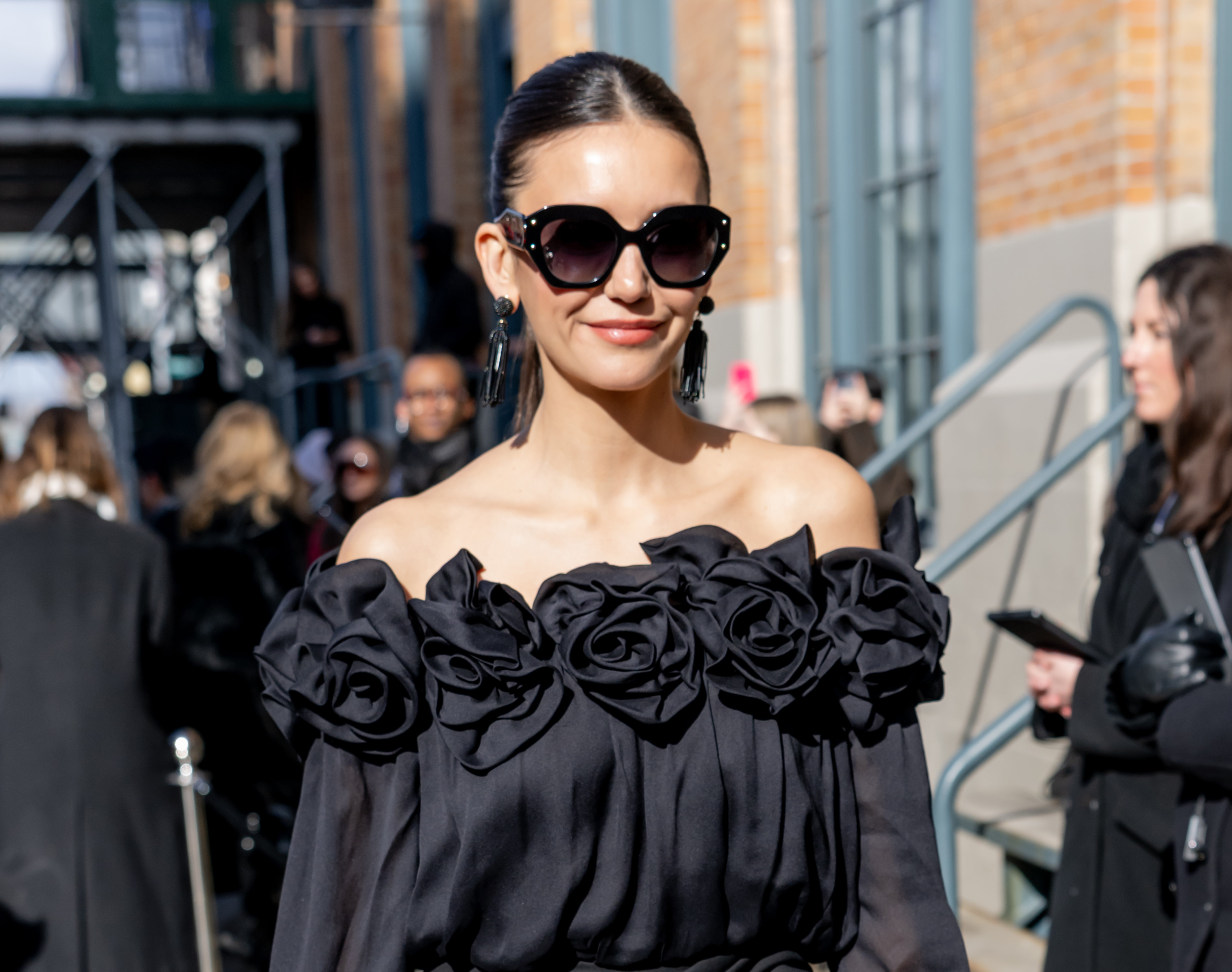 Person in an off-the-shoulder dress adorned with floral details, wearing large sunglasses and earrings, smiling outdoors among people