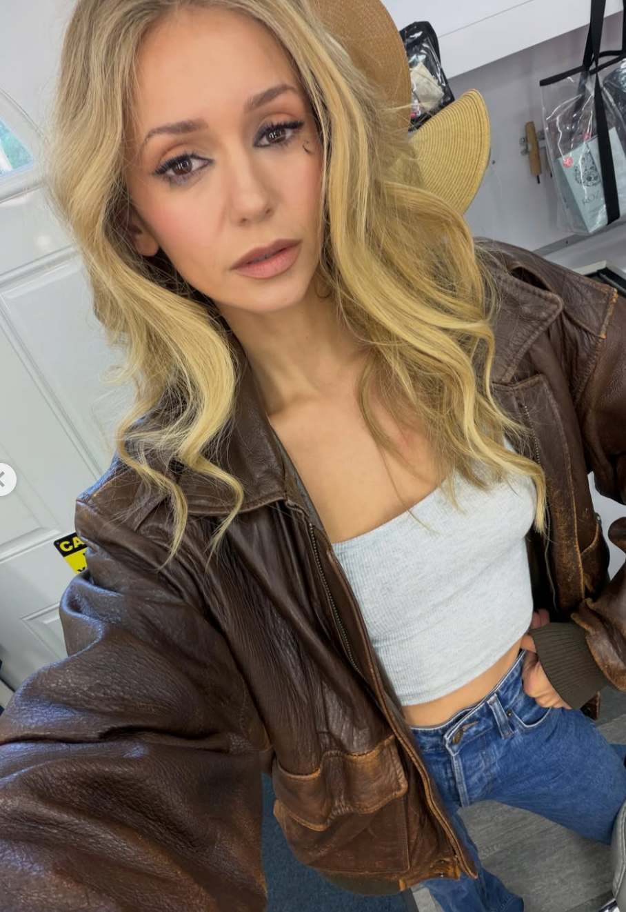 Person with wavy hair wears a large leather jacket, white top, and jeans, posing indoors