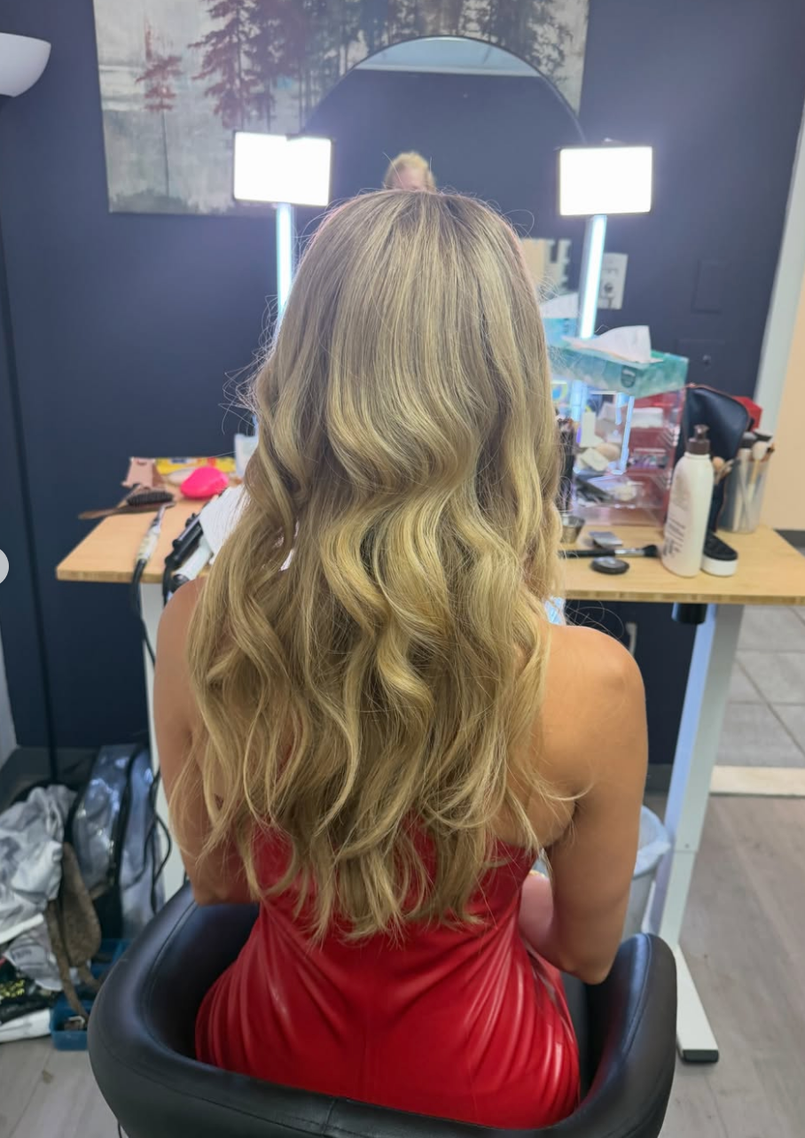 Person with wavy hair seated in front of a makeup vanity, preparing for an event in a fitting red dress